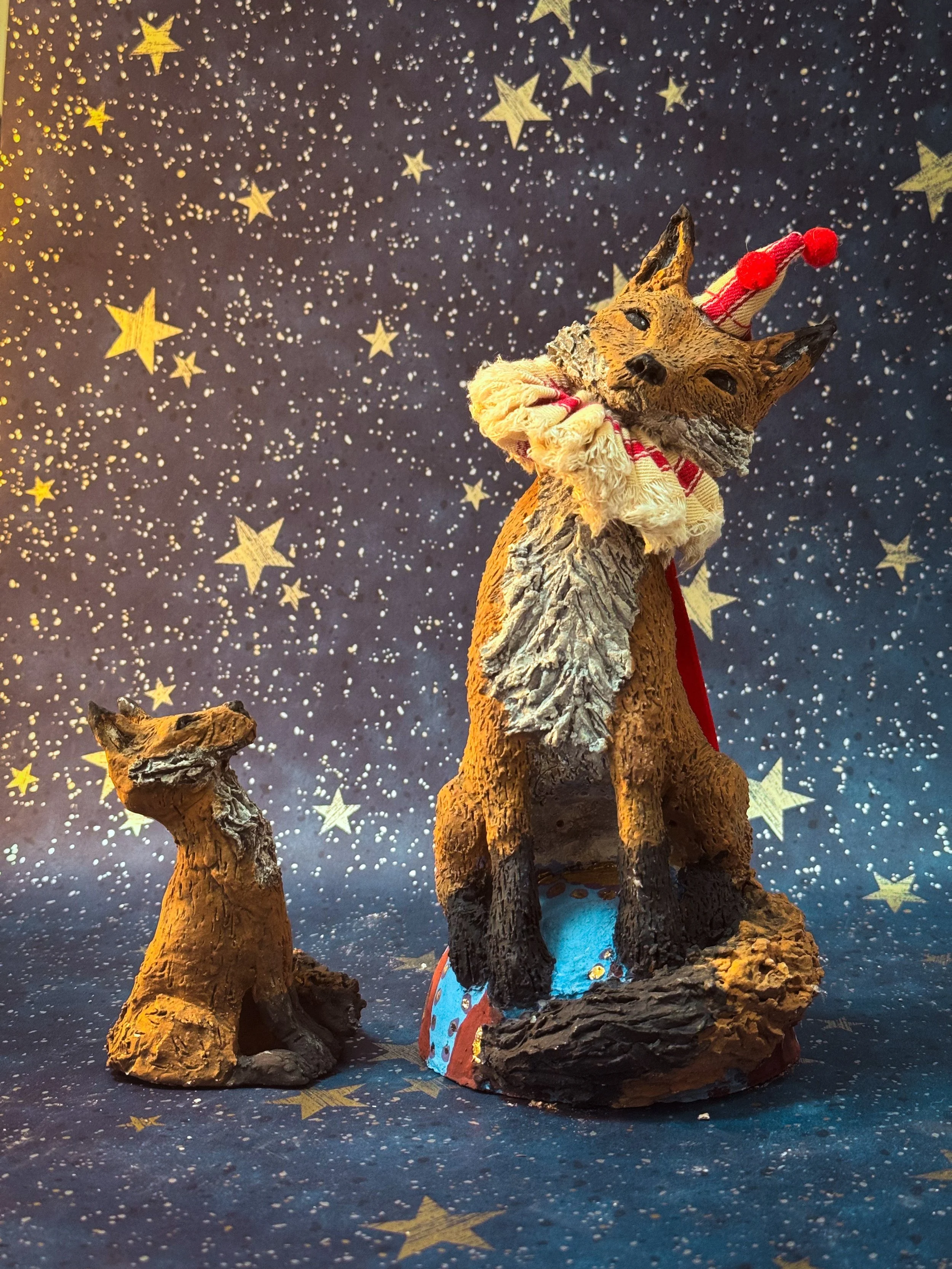 A decorated figurine of a fox wearing a red and white circus  hat and ruffle, sitting on a rocky base with a starry night backdrop.