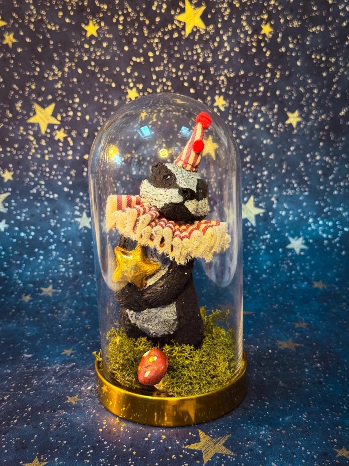 A small decorated sculpture of a badger  wearing a festive clown hat and a ruffled collar inside a glass dome, with a star ornament on its side, set against a starry night background.