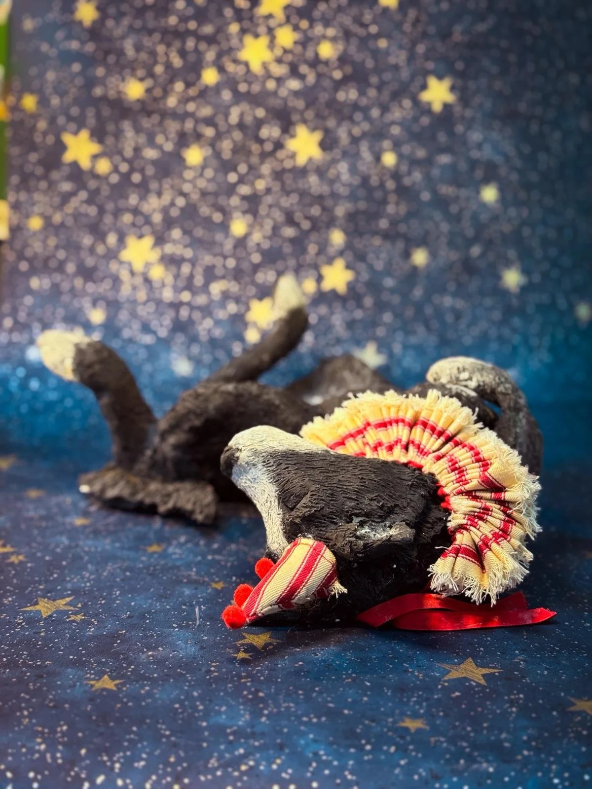 A small black  dog  lying on its back on a starry blue backdrop. It is adorned with a colorful ruffled fabric collar and decorative red pom-poms, with a background decorated with yellow stars.