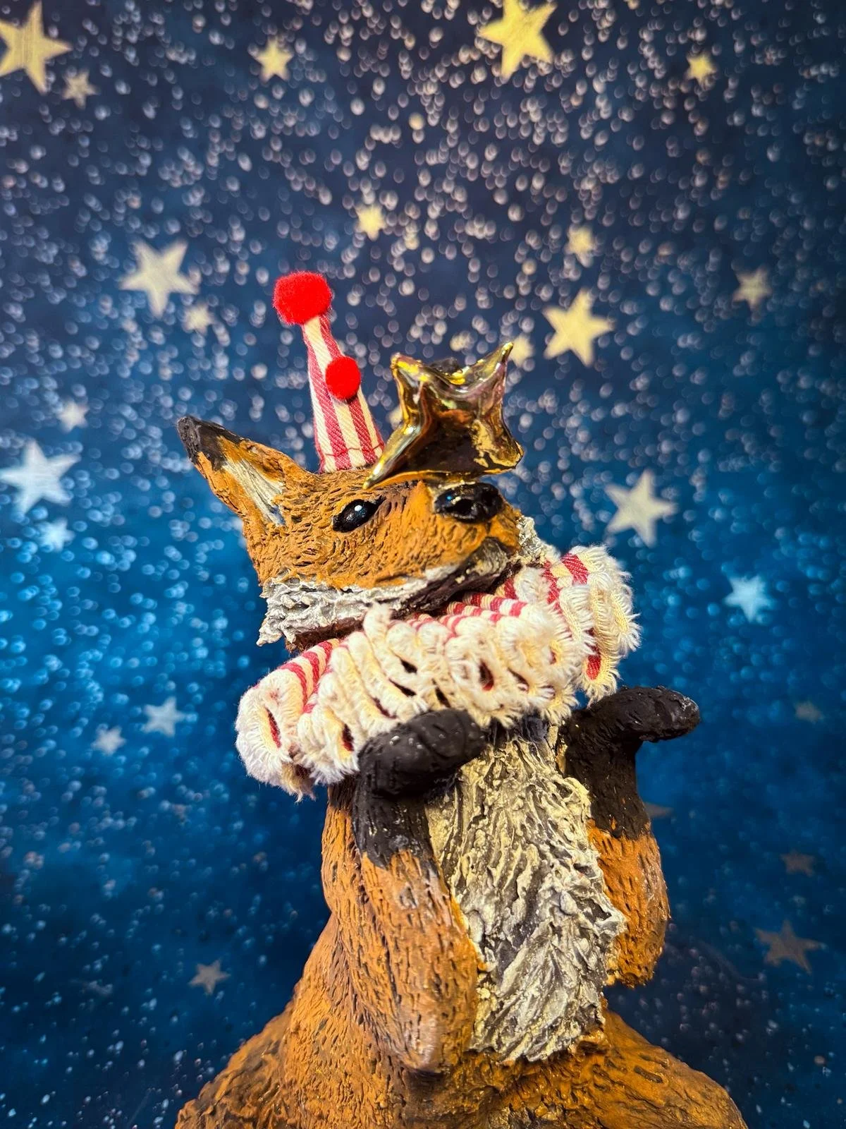 A decorative fox figurine dressed as a clown, with a party hat and a collar, set against a starry night background.