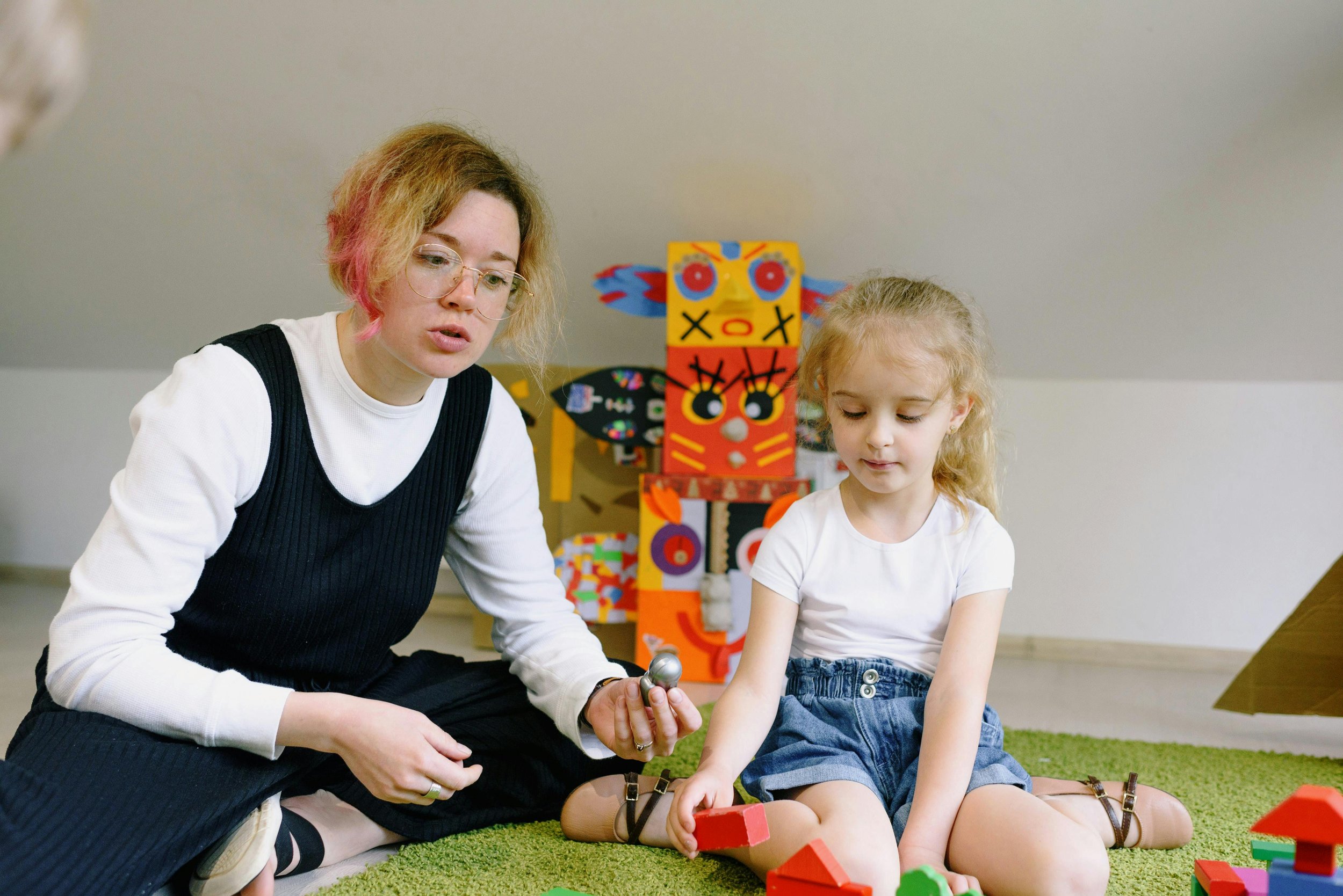 A woman sits on the floor holding a toy and speaks to a child while they are playing with building blocks