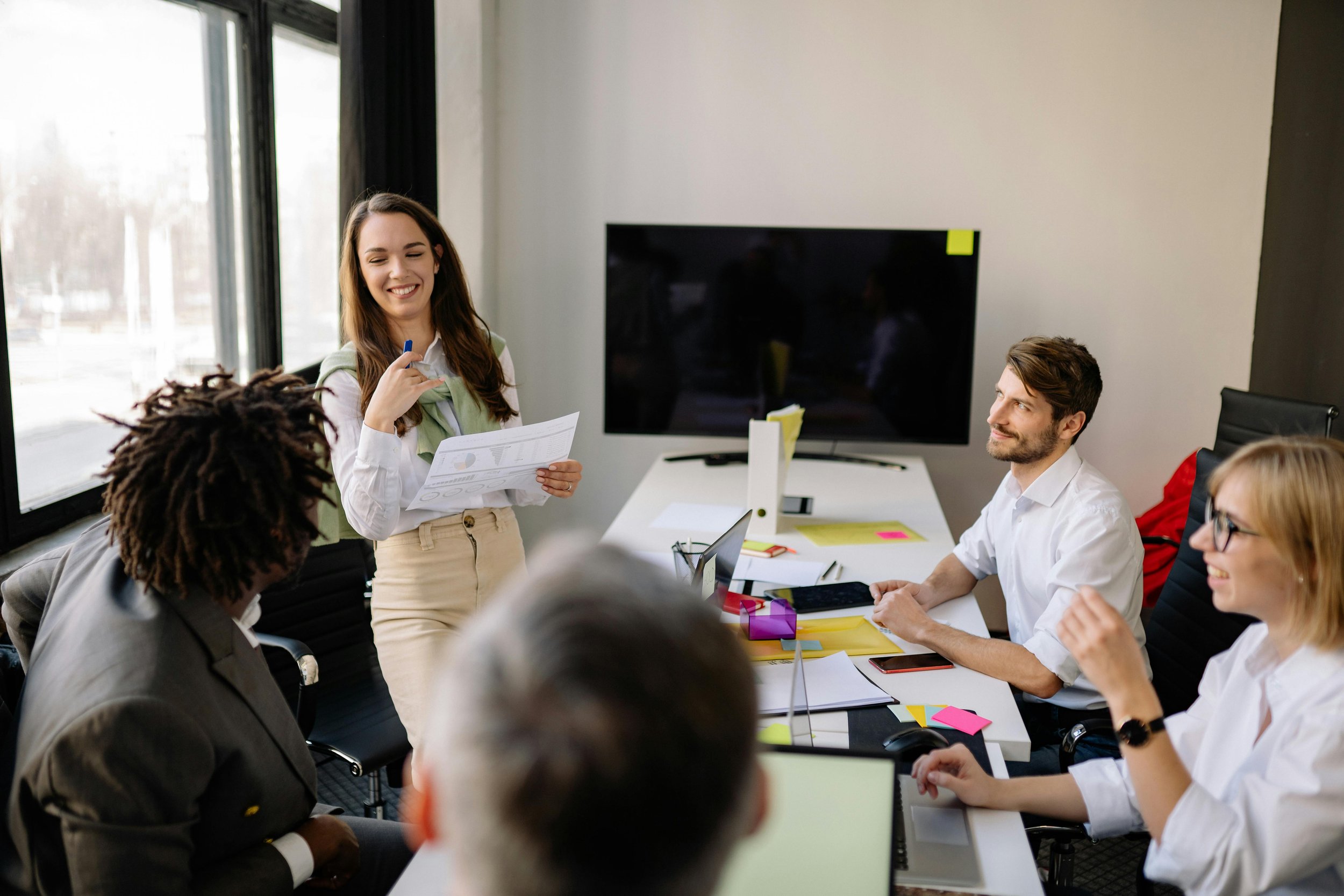 A woman stands at a conference table and leads a presentation to a group of four people that are sitting down