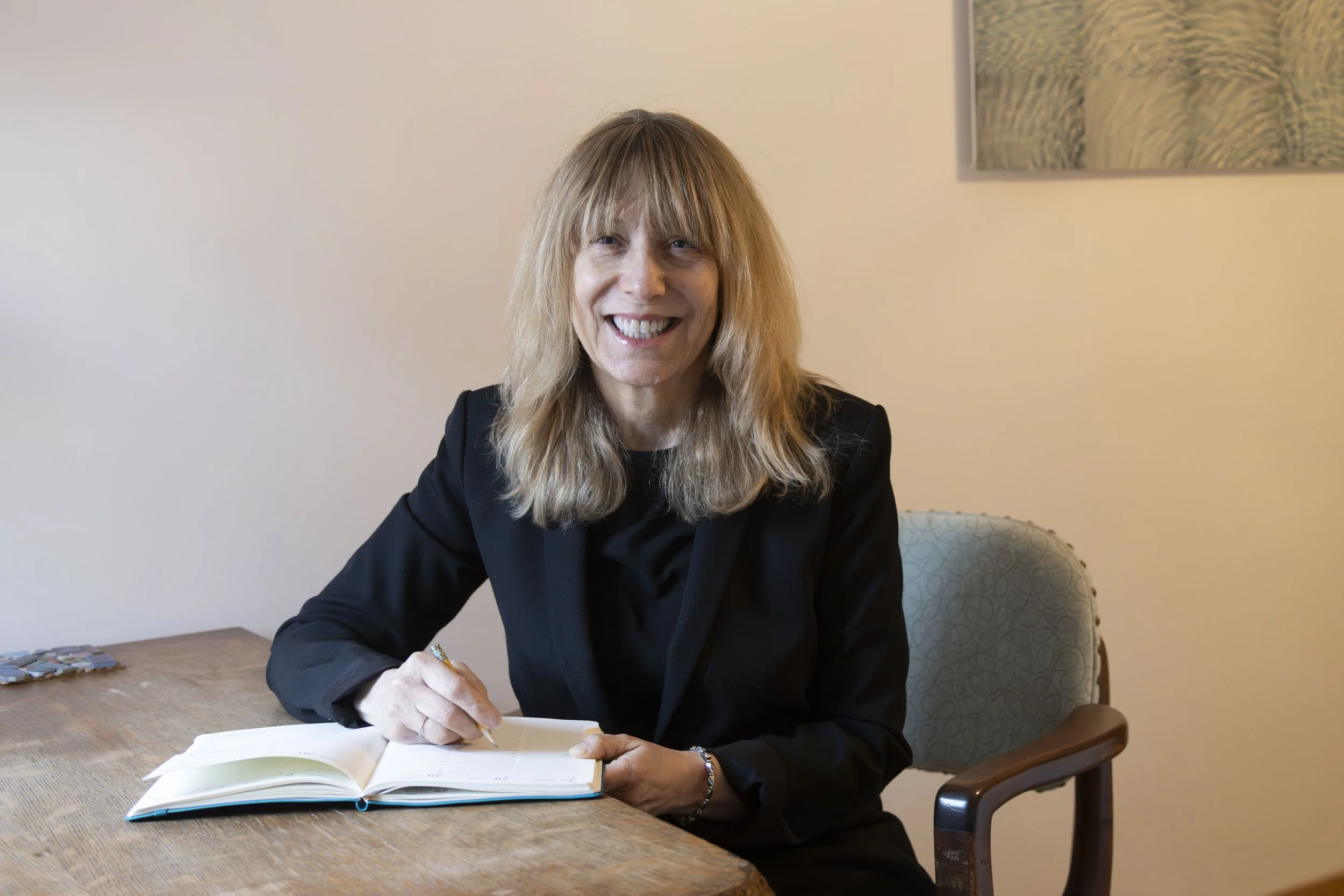 A smiling woman with shoulder-length blonde hair, wearing a black blazer, sitting at a wooden table, signing a book.