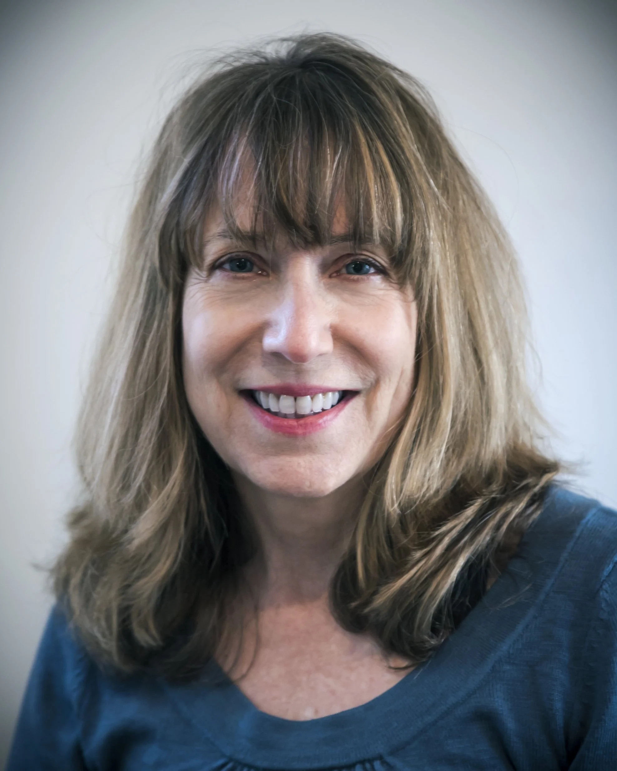 A woman with shoulder-length light brown hair and bangs, smiling, wearing a blue top.