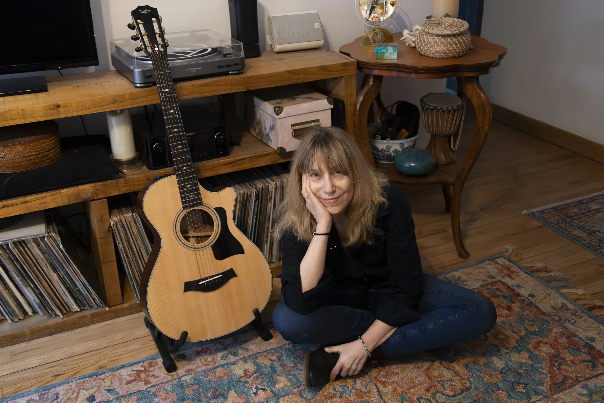 A woman sitting on the floor next to an acoustic guitar, with vinyl records on a shelf behind her, in a cozy living room.