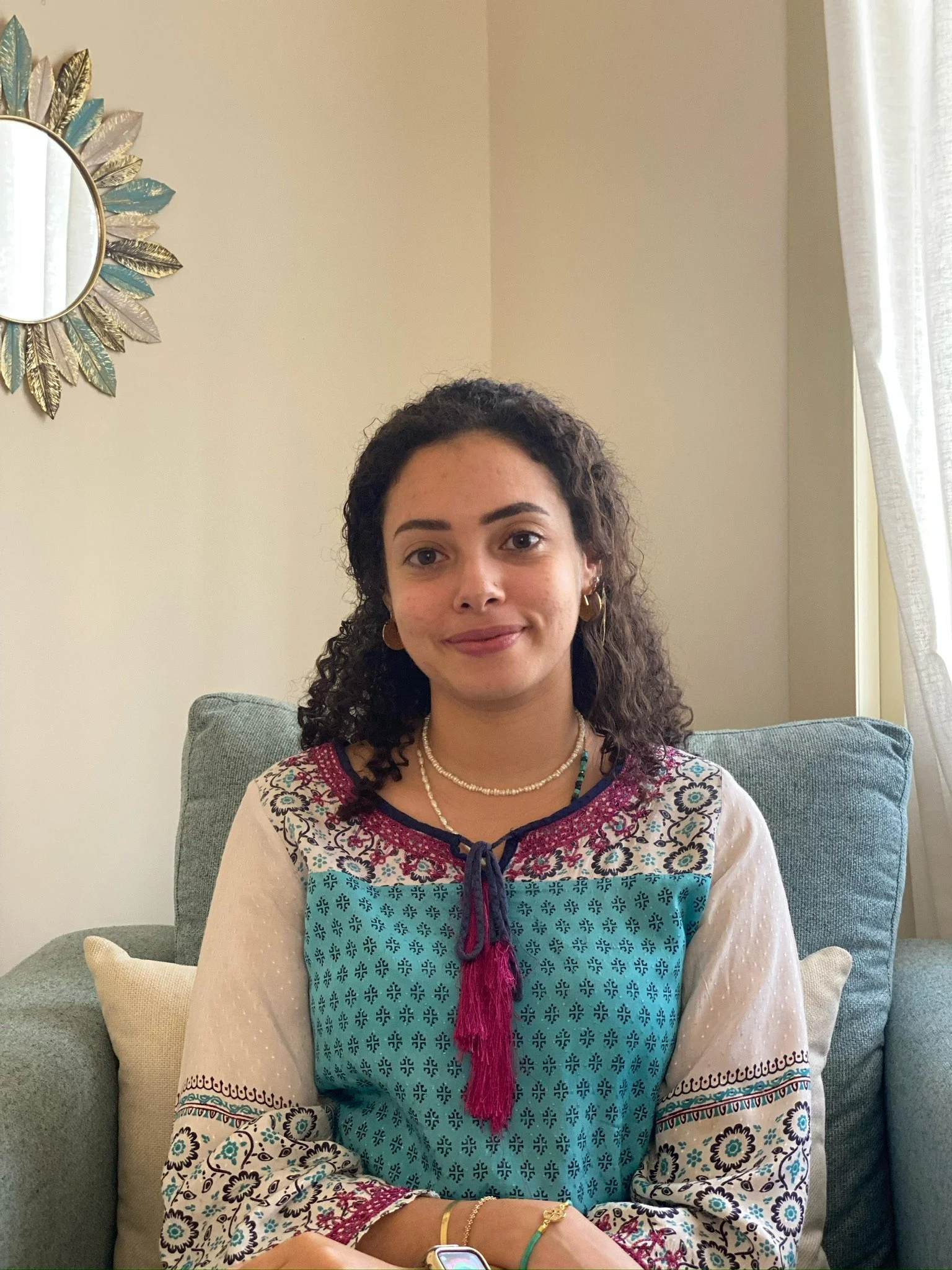 A woman with dark curly hair sitting on a gray sofa with beige and patterned pillows. She is wearing a colorful embroidered top, pearl necklaces, gold earrings, and bracelets. There is a decorative mirror with a leaf design on the wall and a window with curtains in the background.
