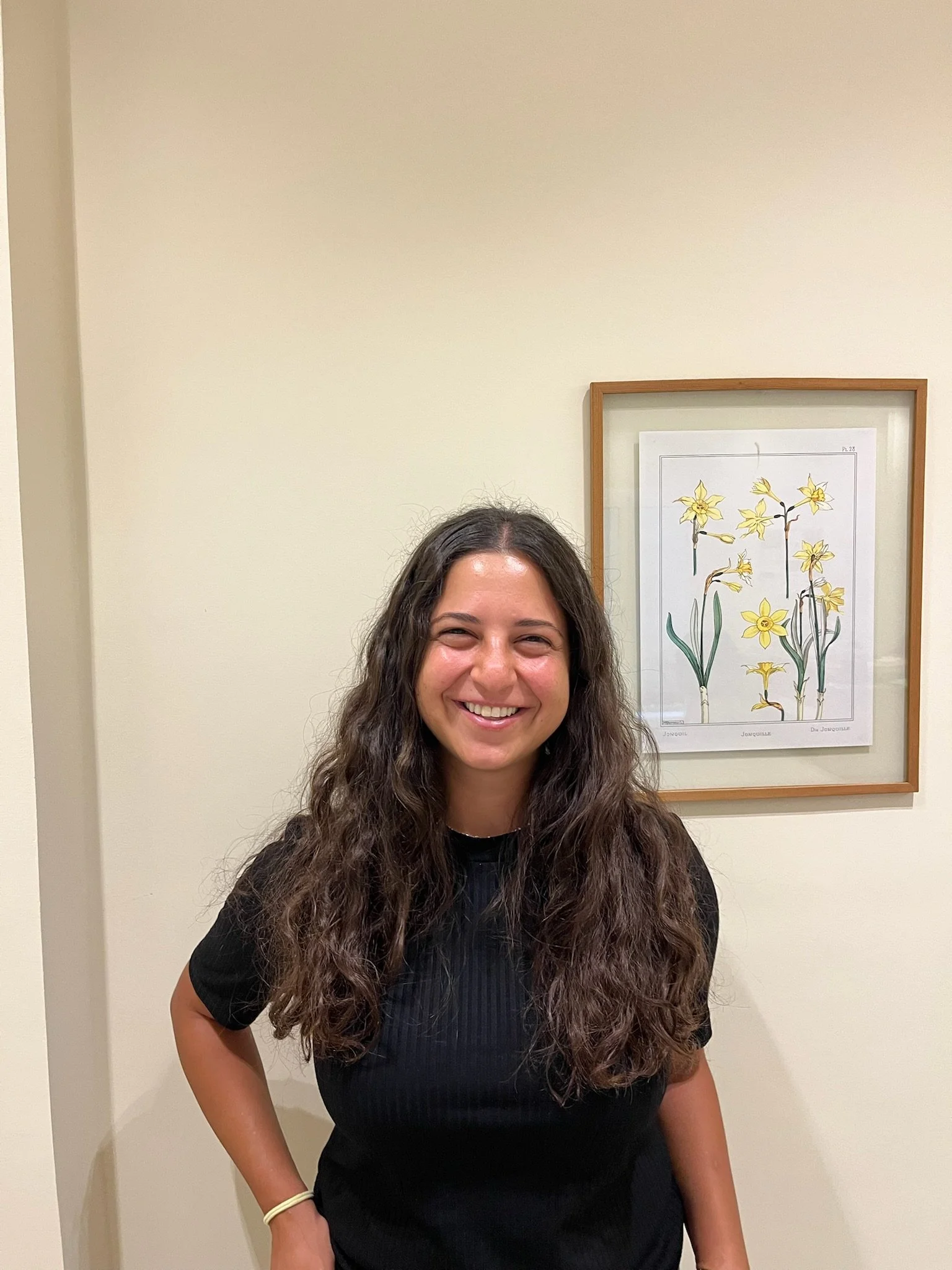 A woman with long, curly brown hair smiling and standing indoors against a beige wall with framed botanical illustration of yellow flowers behind her.