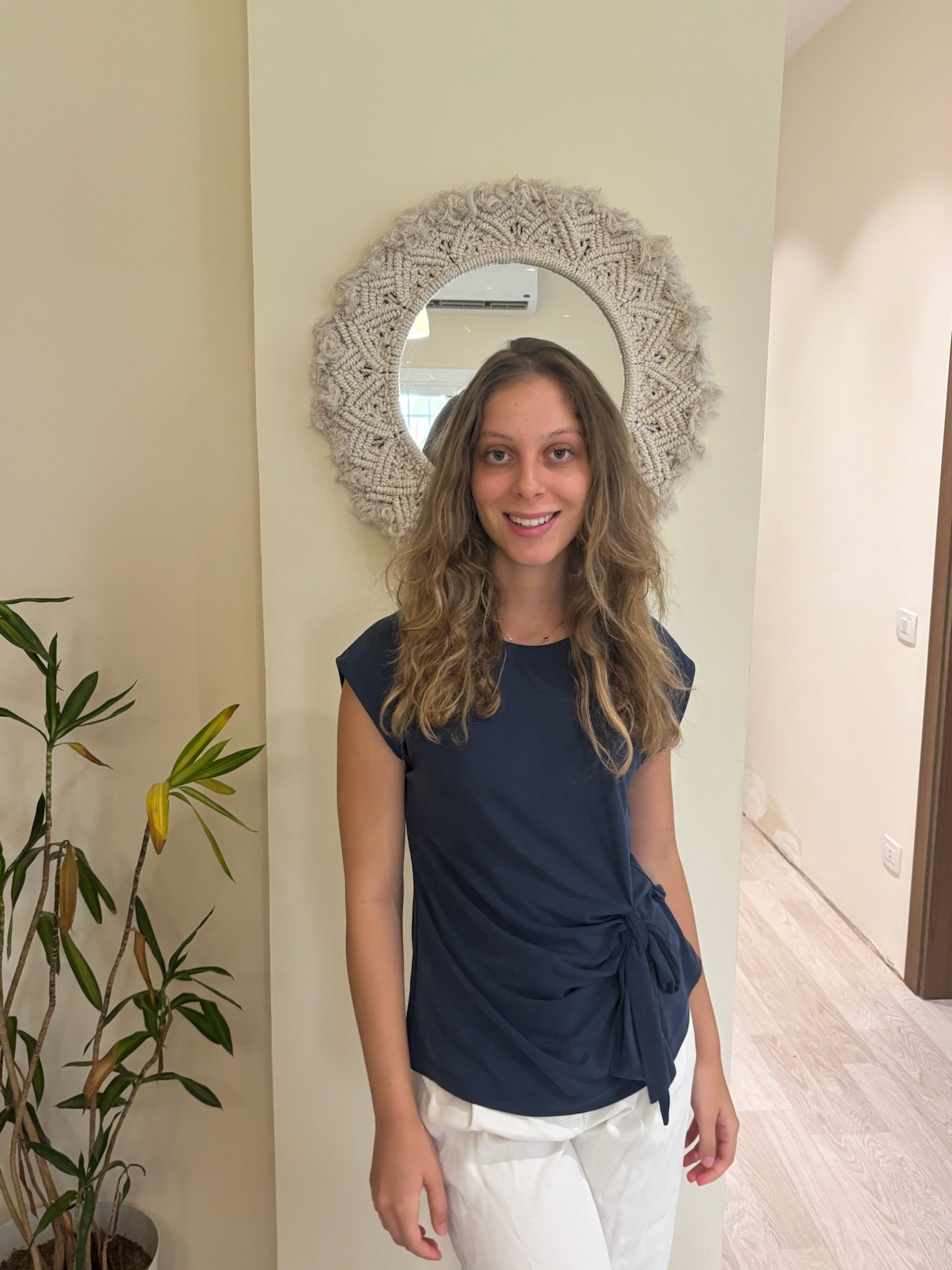 A woman with long blonde curly hair smiling, wearing a navy blue top and white pants, standing in front of a mirror with a knitted frame, in an indoor setting with cream-colored walls and a potted plant nearby.