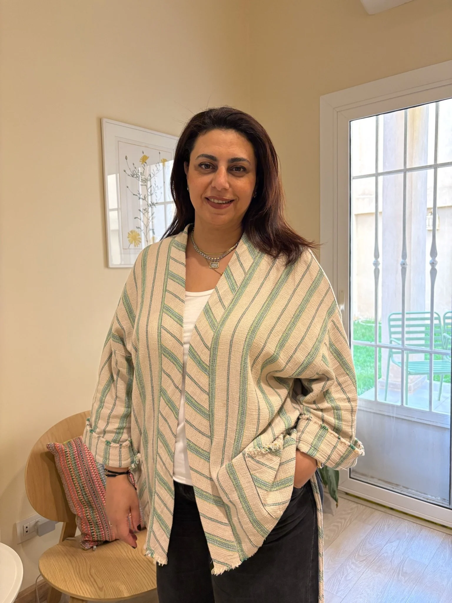 A woman standing indoors near a glass door with an outdoor patio visible. She has shoulder-length dark hair, wearing a striped beige and white cardigan, white top, and black pants. She is smiling, with her right hand in her pocket.