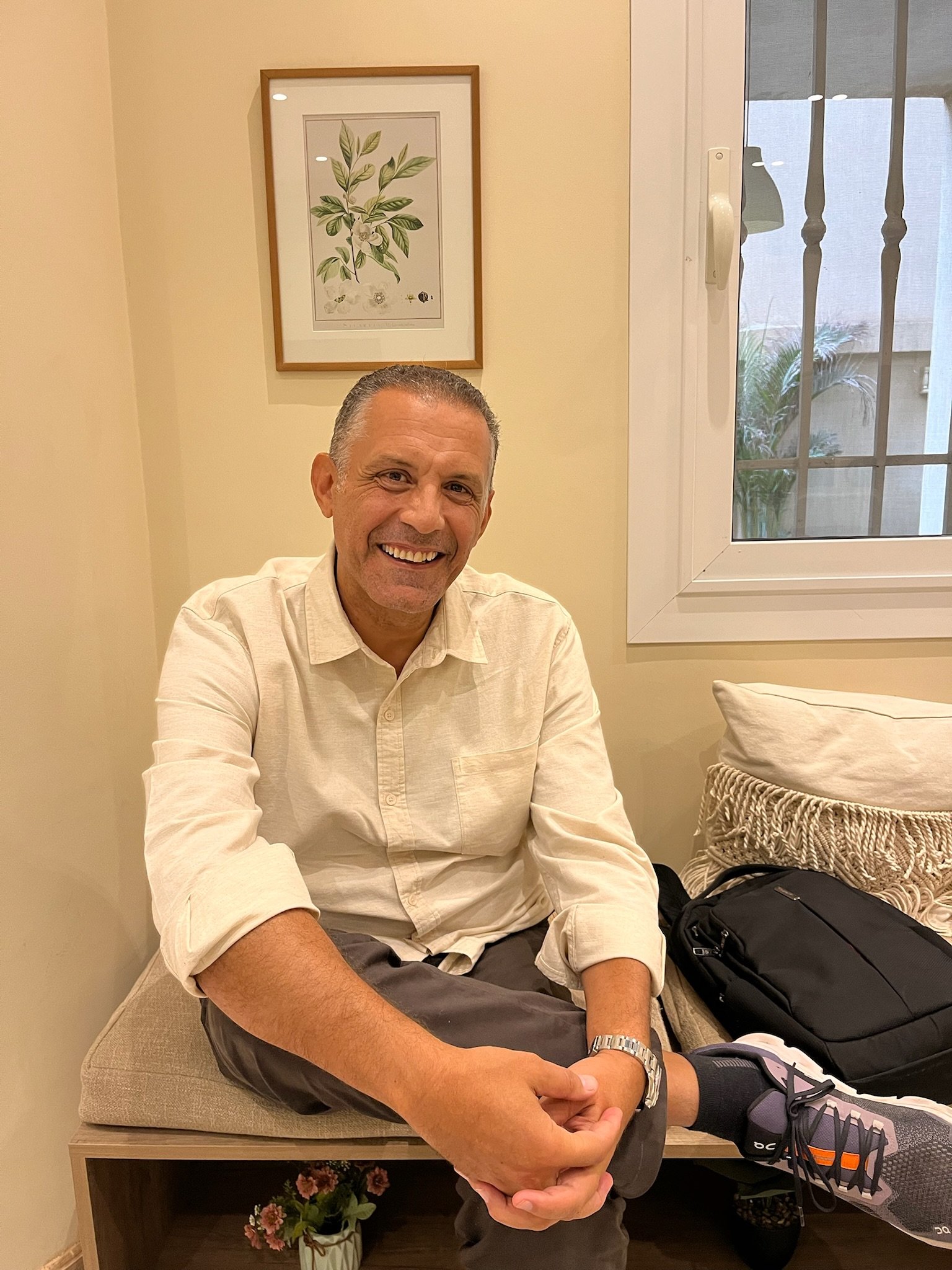 A smiling man sitting on a bench indoors, wearing a cream-colored shirt, gray pants, and sneakers, with a backpack and a pillow nearby, a window behind him, and framed botanical artwork on the wall.