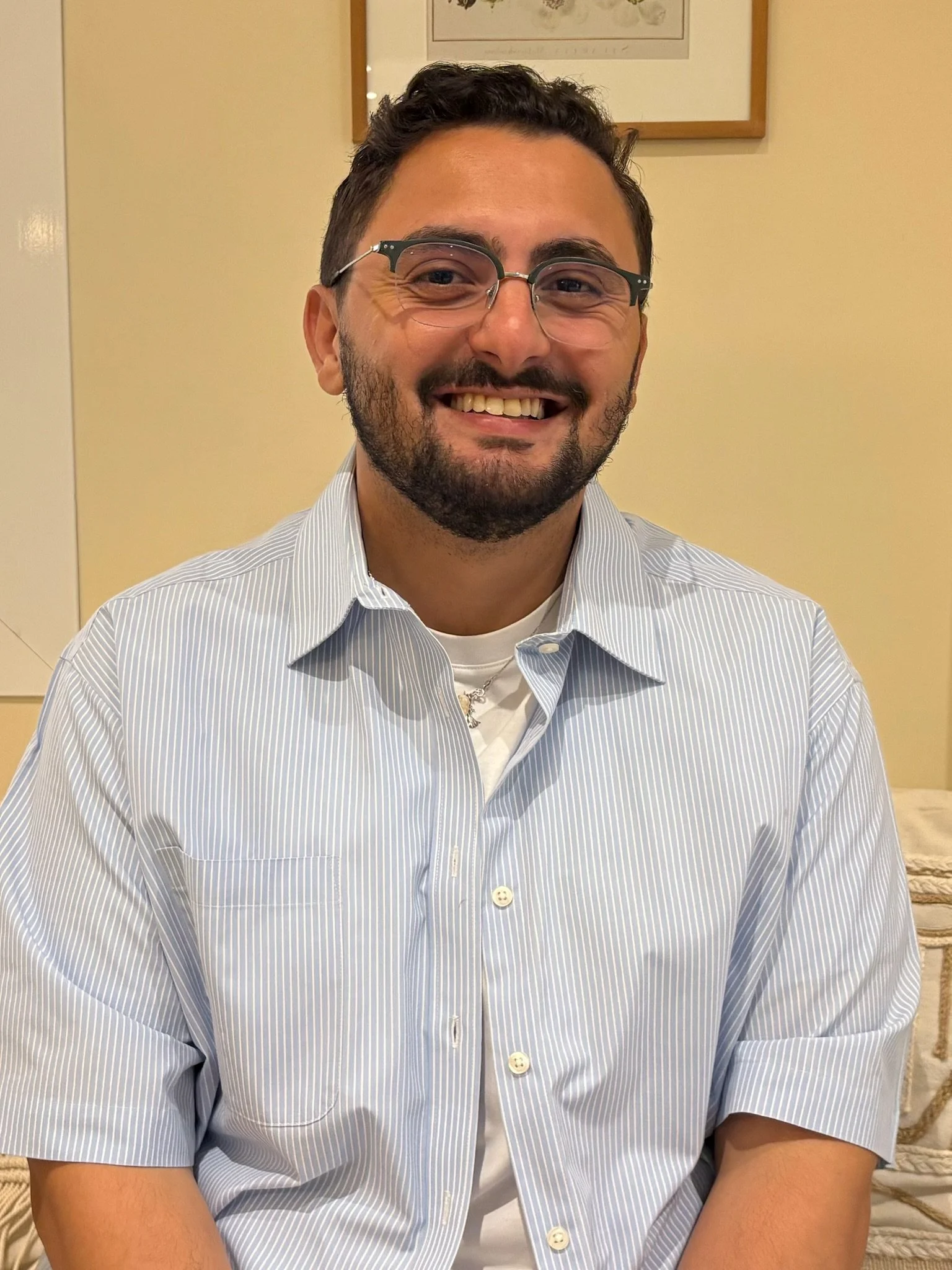 A smiling man with glasses and a beard, wearing a light blue striped short-sleeve shirt, seated indoors with artwork on the wall behind him.