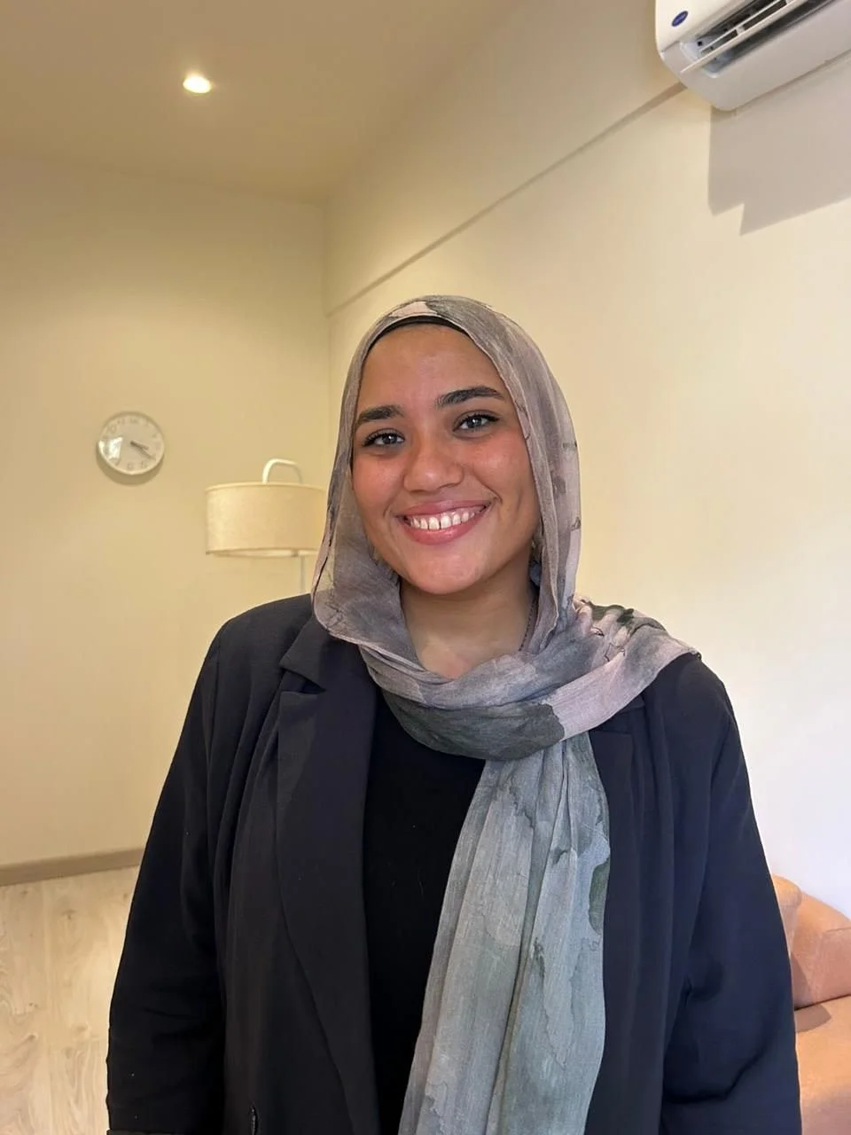 A young woman wearing a grey headscarf and black blazer smiling indoors.
