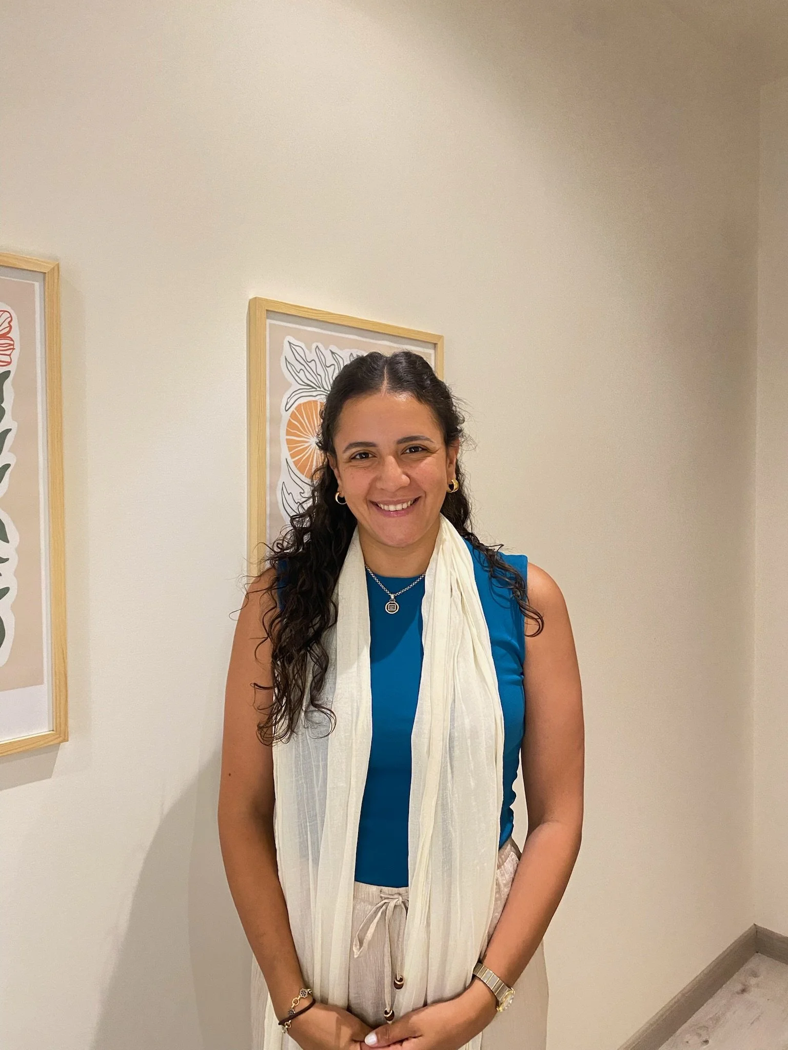 A woman with dark, curly hair wearing a blue sleeveless top, a white scarf, beige pants, and jewelry, standing in an art gallery with framed artwork on the wall behind her, smiling at the camera.