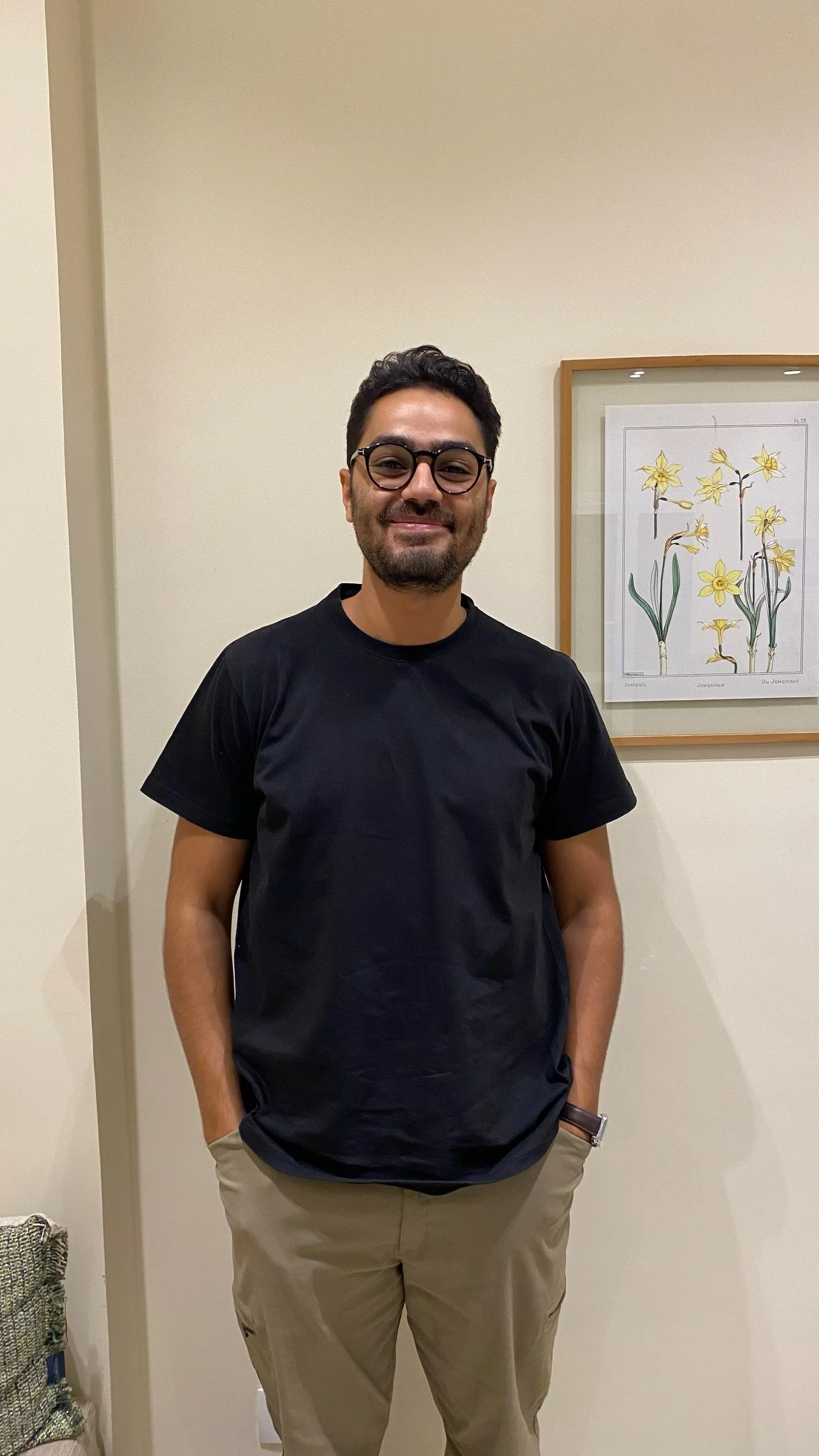 A man with dark hair, wearing glasses, a black t-shirt, and khaki pants, standing indoors against a beige wall next to framed botanical artwork.