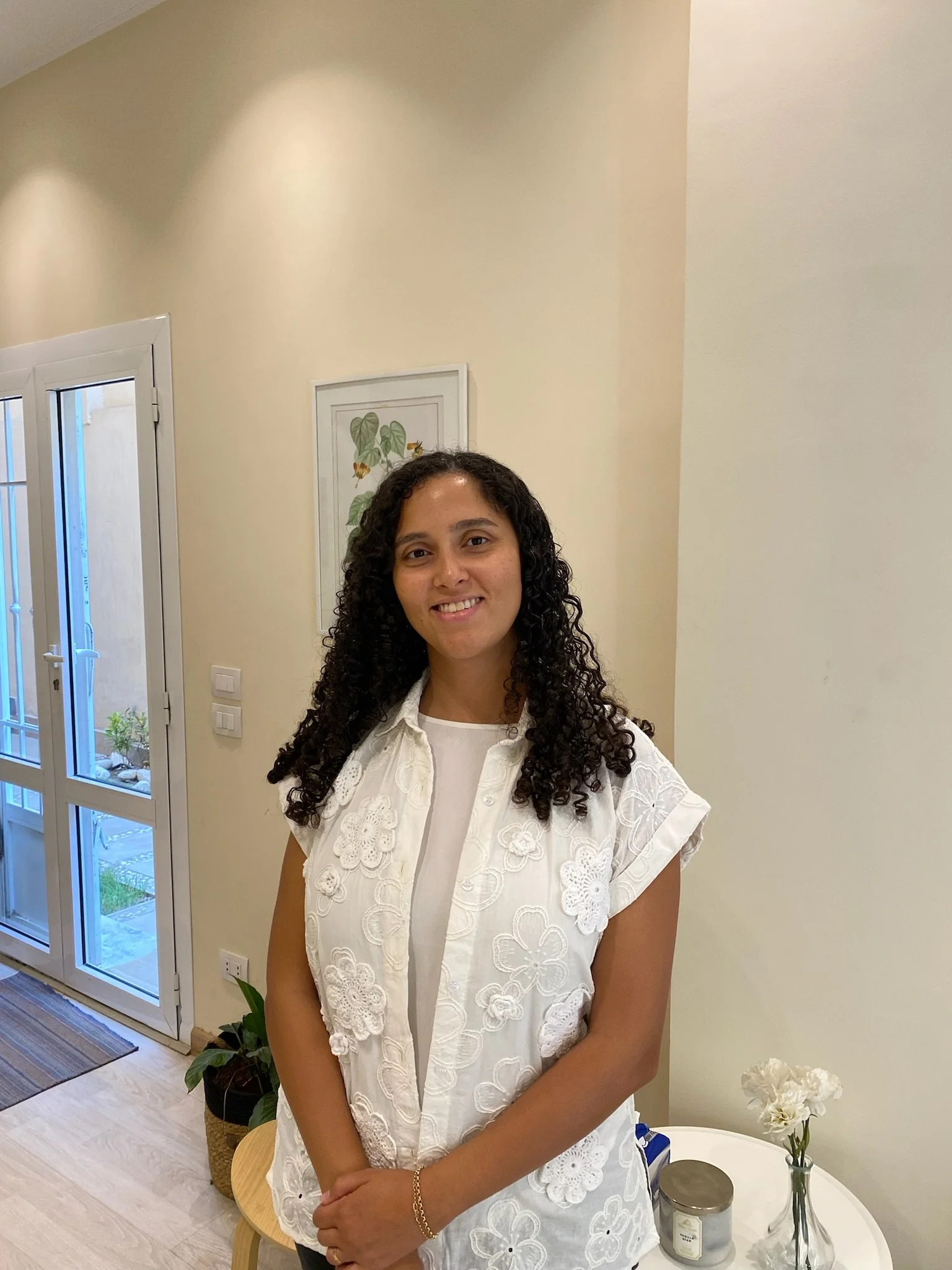 A woman with curly dark hair wearing a white embroidered sleeveless top, standing indoors near a white table with a candle and flowers, in a room with beige walls and a plant in the background.