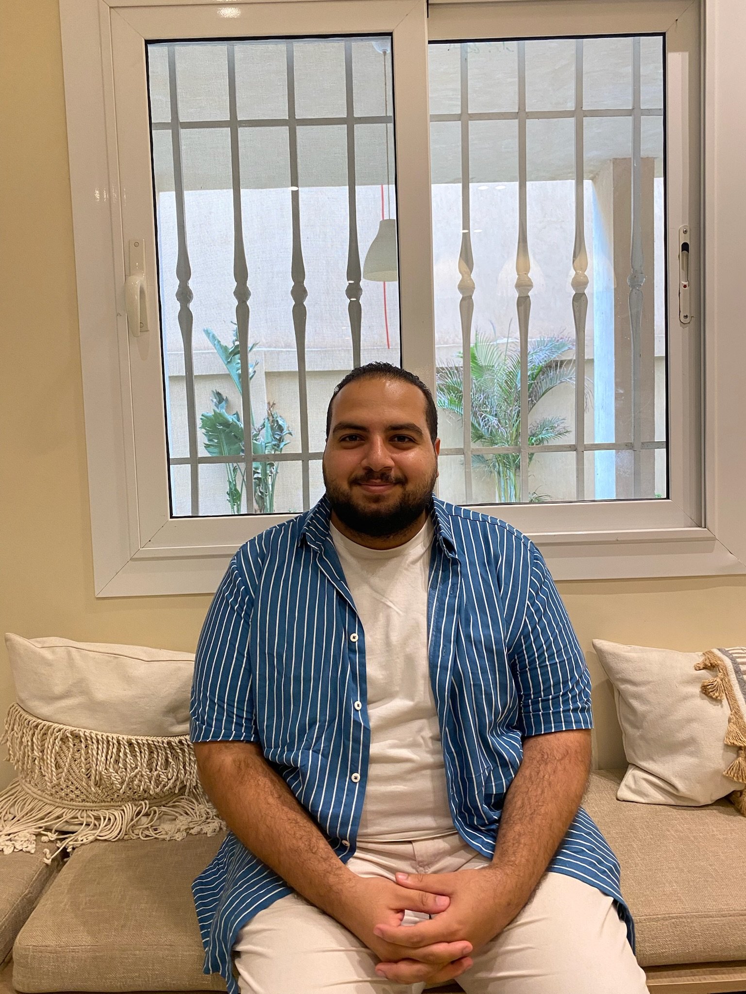 A man sitting on a beige sofa, smiling, in front of a large window with white frame and metal bars, with a view of plants outside.