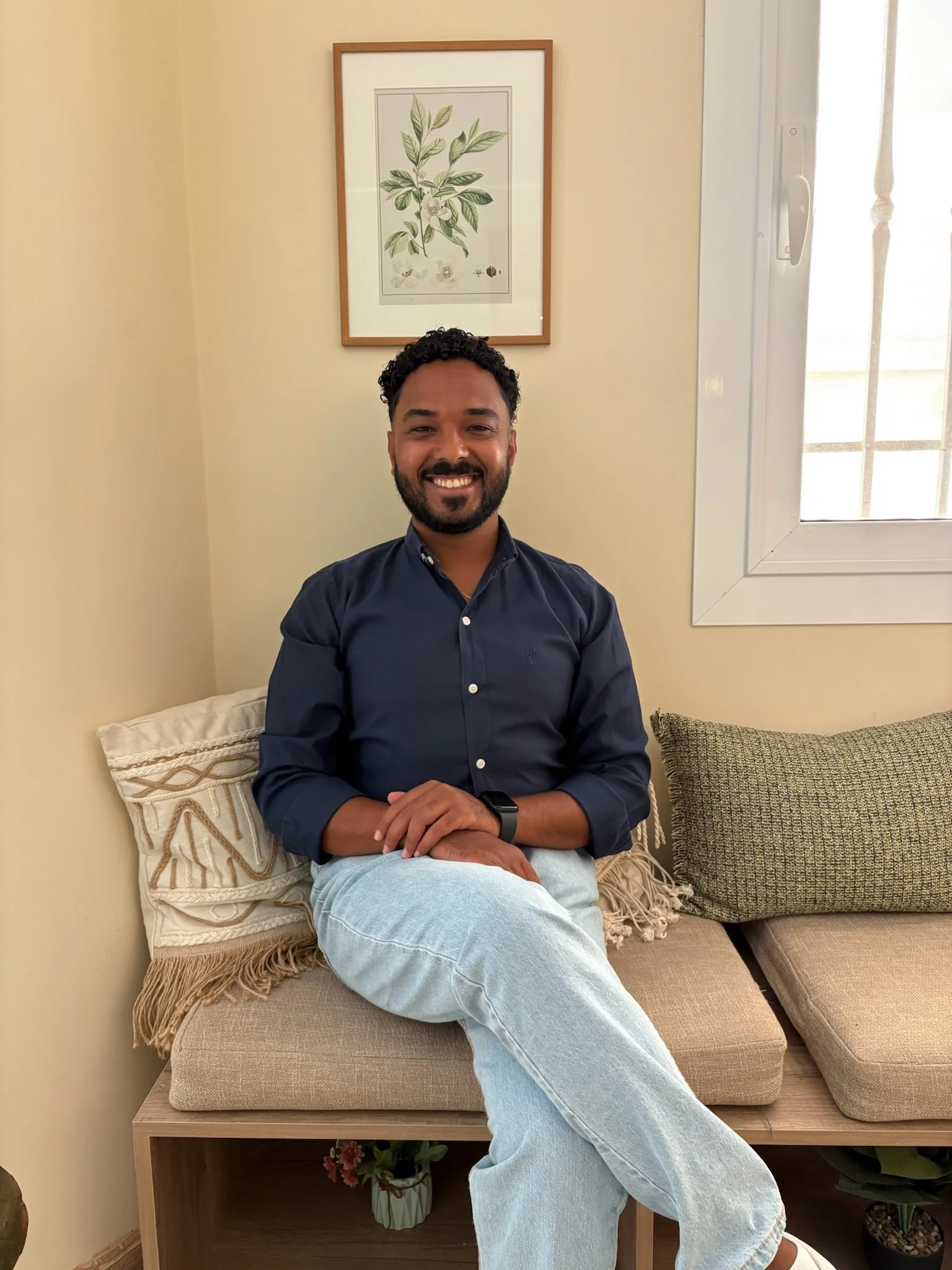A man with curly dark hair and a beard sitting on a beige cushioned bench, smiling at the camera. He is wearing a navy blue button-up shirt, light blue jeans, and a black wristwatch. The background features a framed botanical print on a beige wall, a window with white framing, and some decorative pillows and plants.