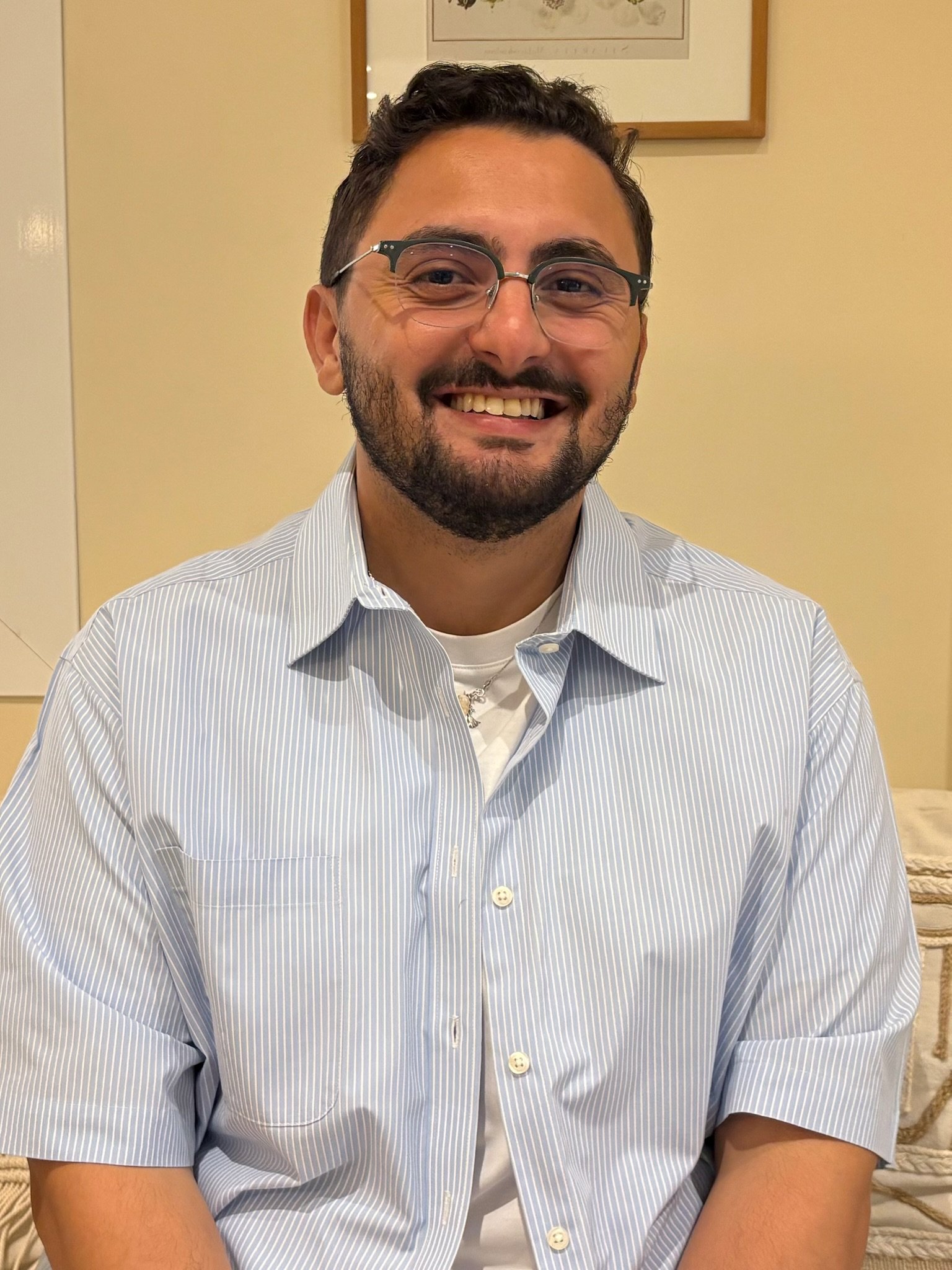 A man with glasses and a beard is smiling, wearing a light blue striped shirt, sitting indoors against a beige wall with a framed artwork in the background.