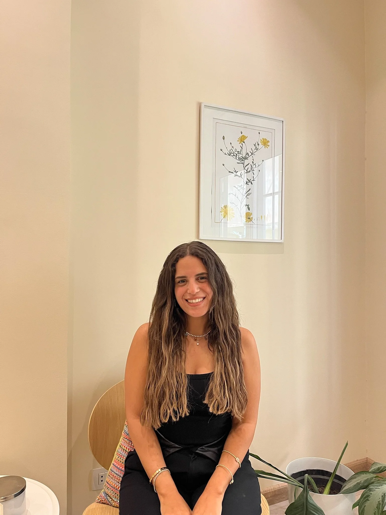 A woman with long wavy hair sitting on a wooden chair, smiling at the camera, next to a potted plant in a white pot on the floor, with a framed floral artwork on the cream-colored wall behind her.
