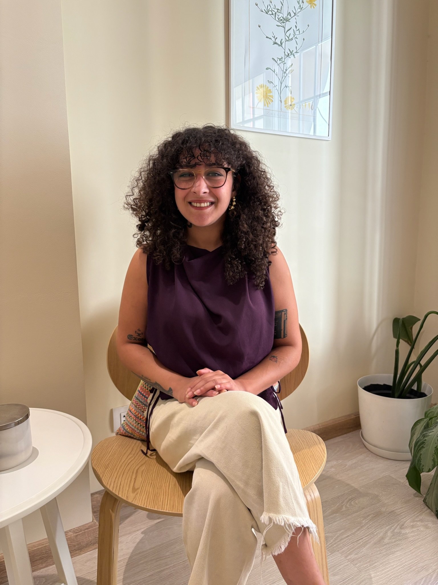 A woman with curly dark hair and glasses smiling, sitting on a wooden chair in a room with light-colored walls, a window with floral designs, and potted plants.