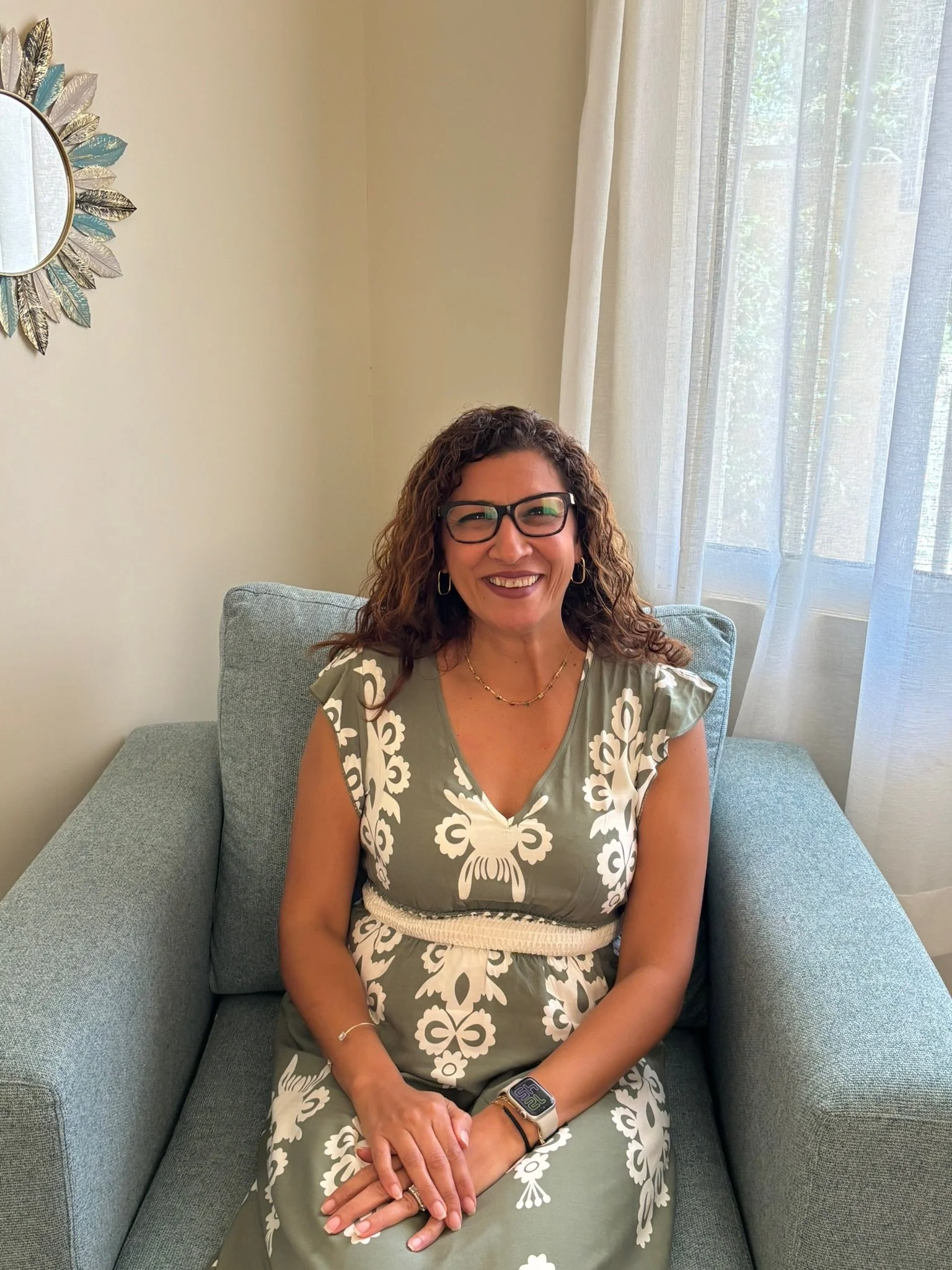 A woman with curly brown hair, wearing glasses, a green dress with white floral patterns, and jewelry, sitting on a light blue sofa in front of a cream wall and a window with white curtains.