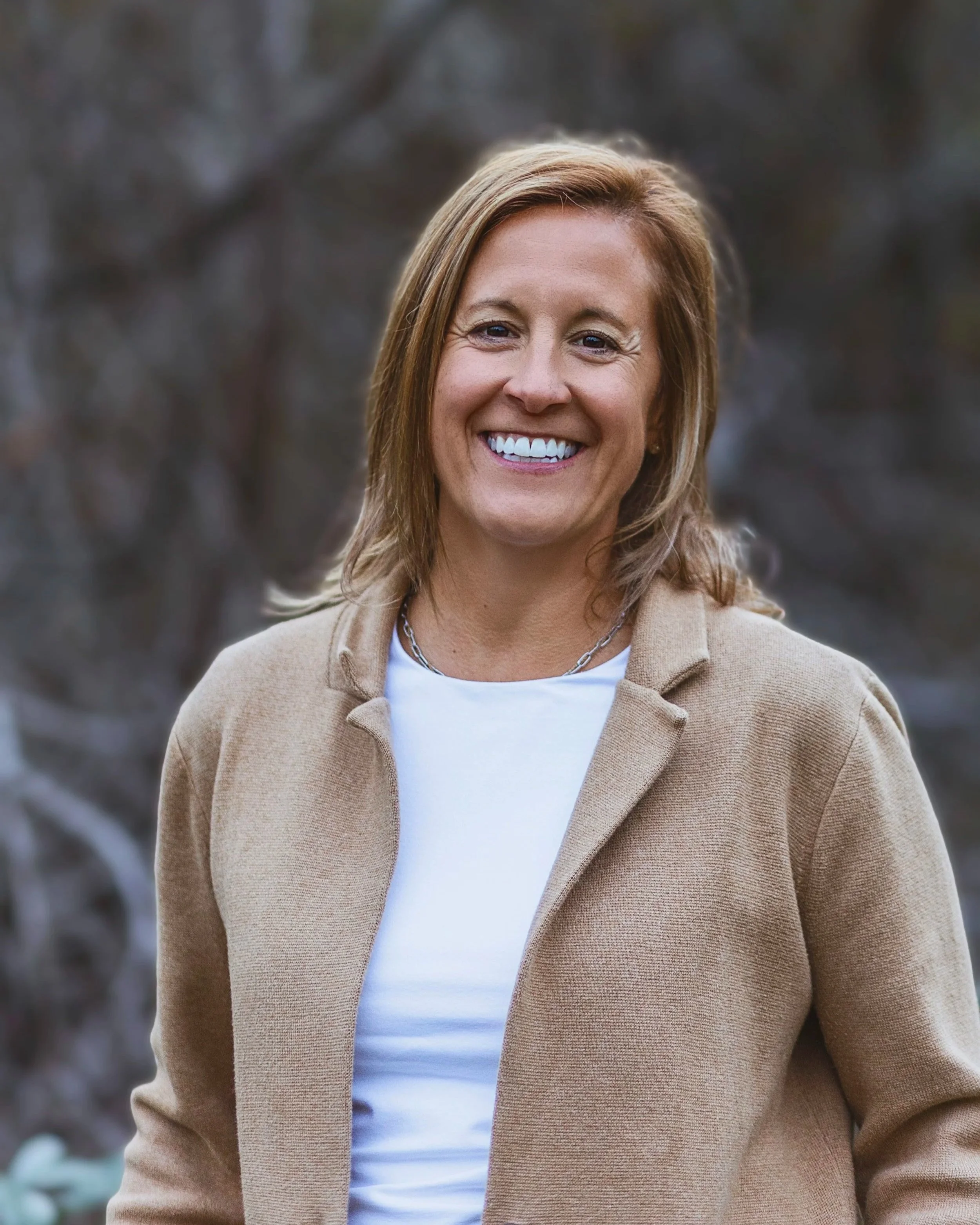 A smiling woman with shoulder-length light brown hair outdoors, wearing a beige jacket over a white top, with a blurred natural background.