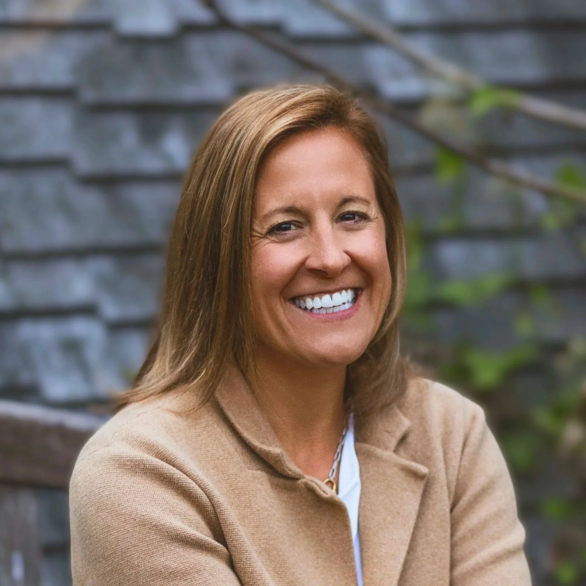 A woman with shoulder-length light brown hair smiling outdoors with a blurred dark wooden fence and green foliage in the background.