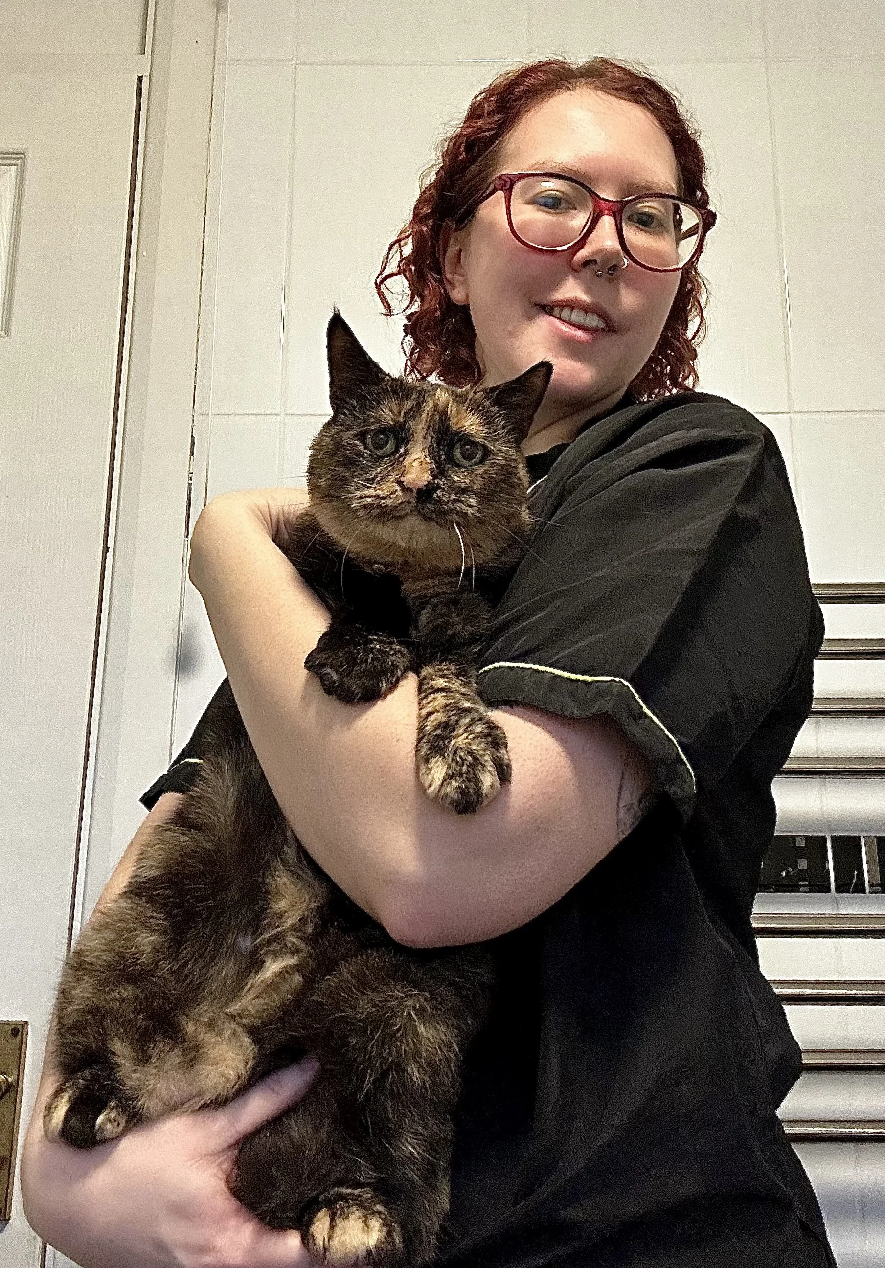 A young woman with red hair, glasses, and a nose piercing holding a tortoiseshell cat in her arms.