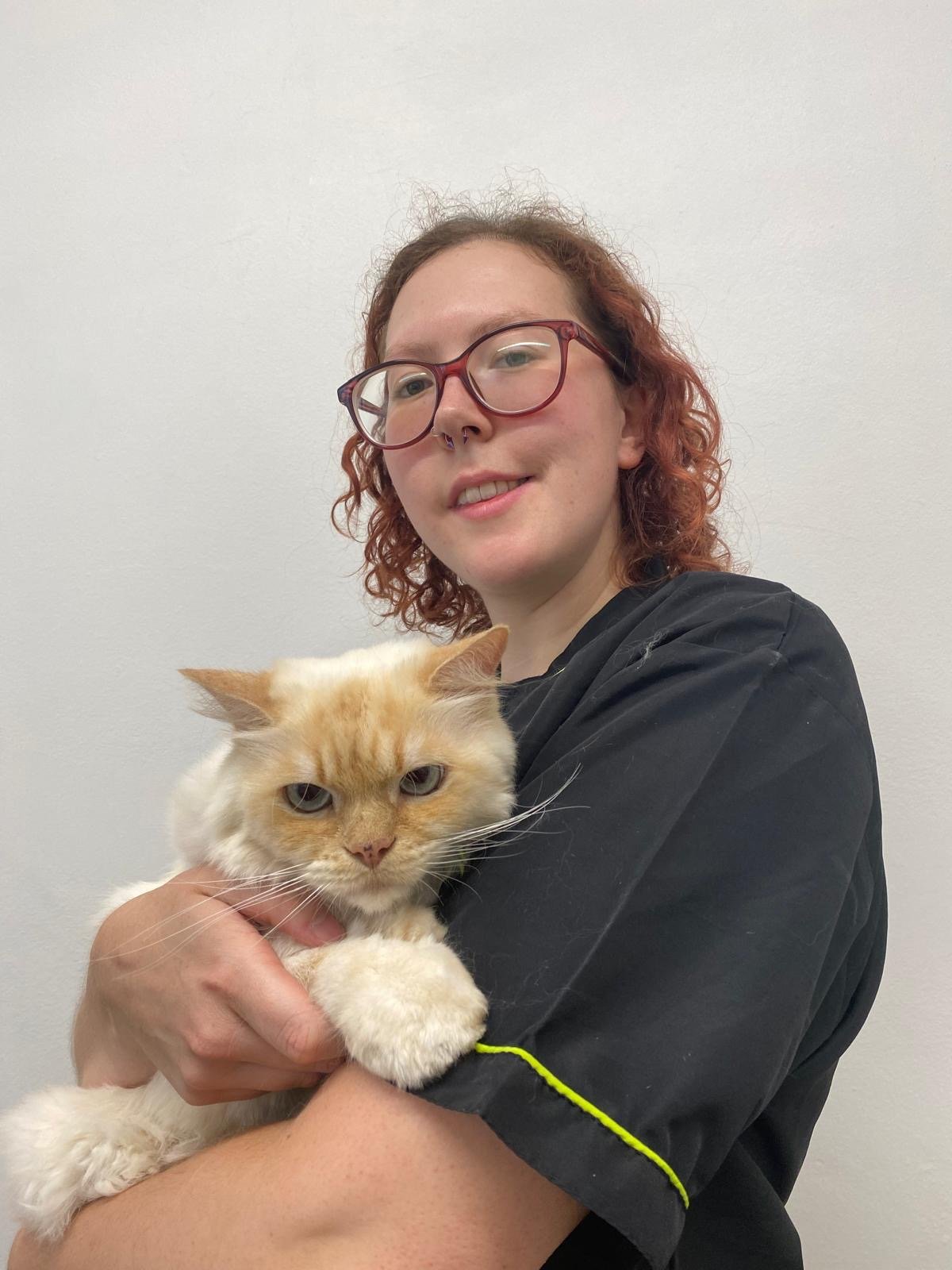 A person with curly red hair, glasses, and a septum piercing smiling while holding an orange and white fluffy cat in front of a plain white wall.