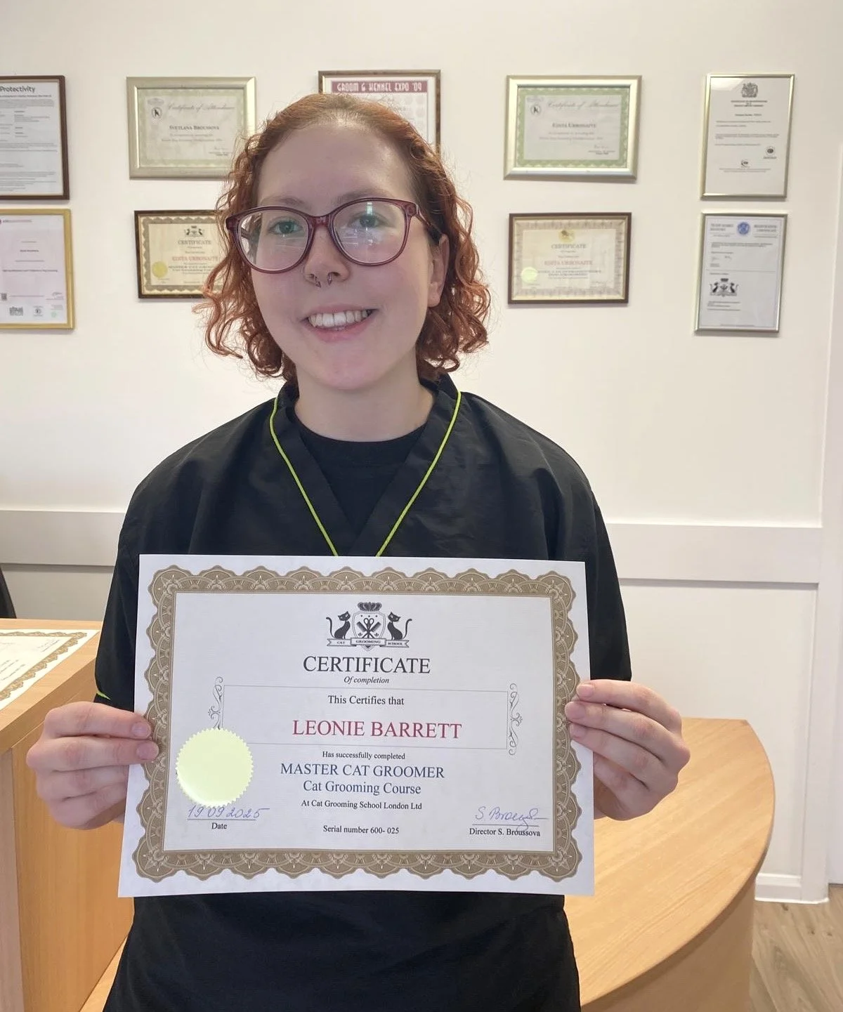 A young woman with curly red hair and glasses smiling while holding a certificate of completion for a cat grooming course.