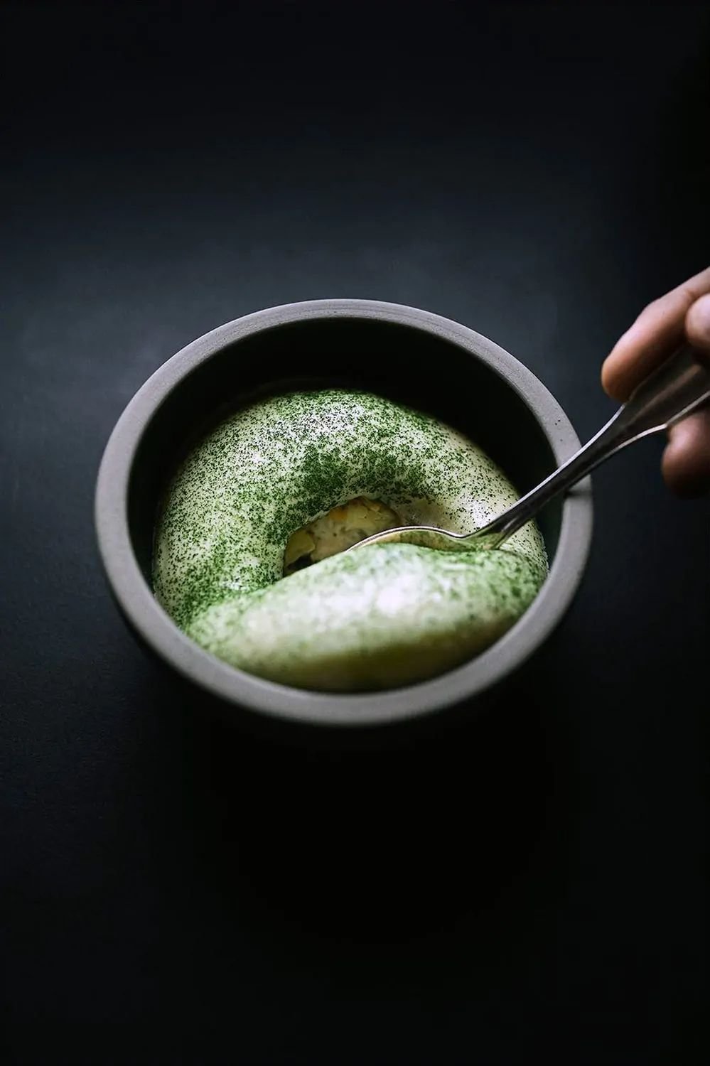 Un cuenco con helado de matcha y una bola de vainilla, acompañado de polvo de matcha espolvoreado encima, y una mano que sostiene una cuchara.