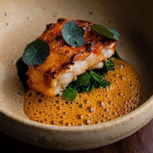 Porción de pescado a la parrilla sobre una espuma anaranjada con vegetales verdes y hojas decorativas