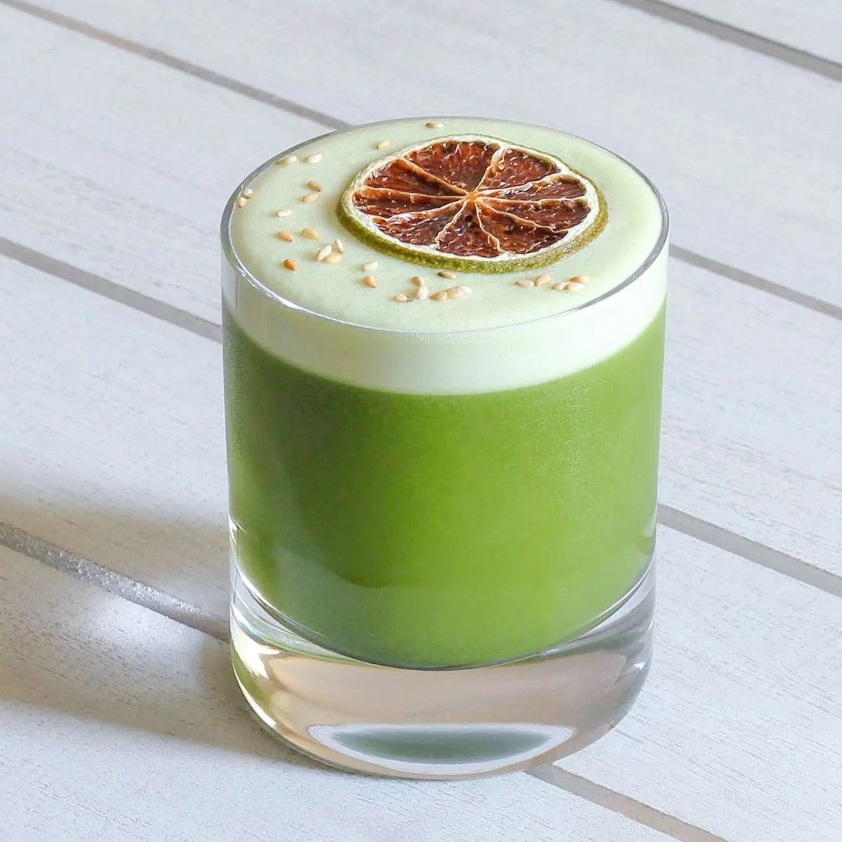 Vaso con bebida verde decorada con rodaja de limón seco y semillas en la parte superior sobre mesa blanca.