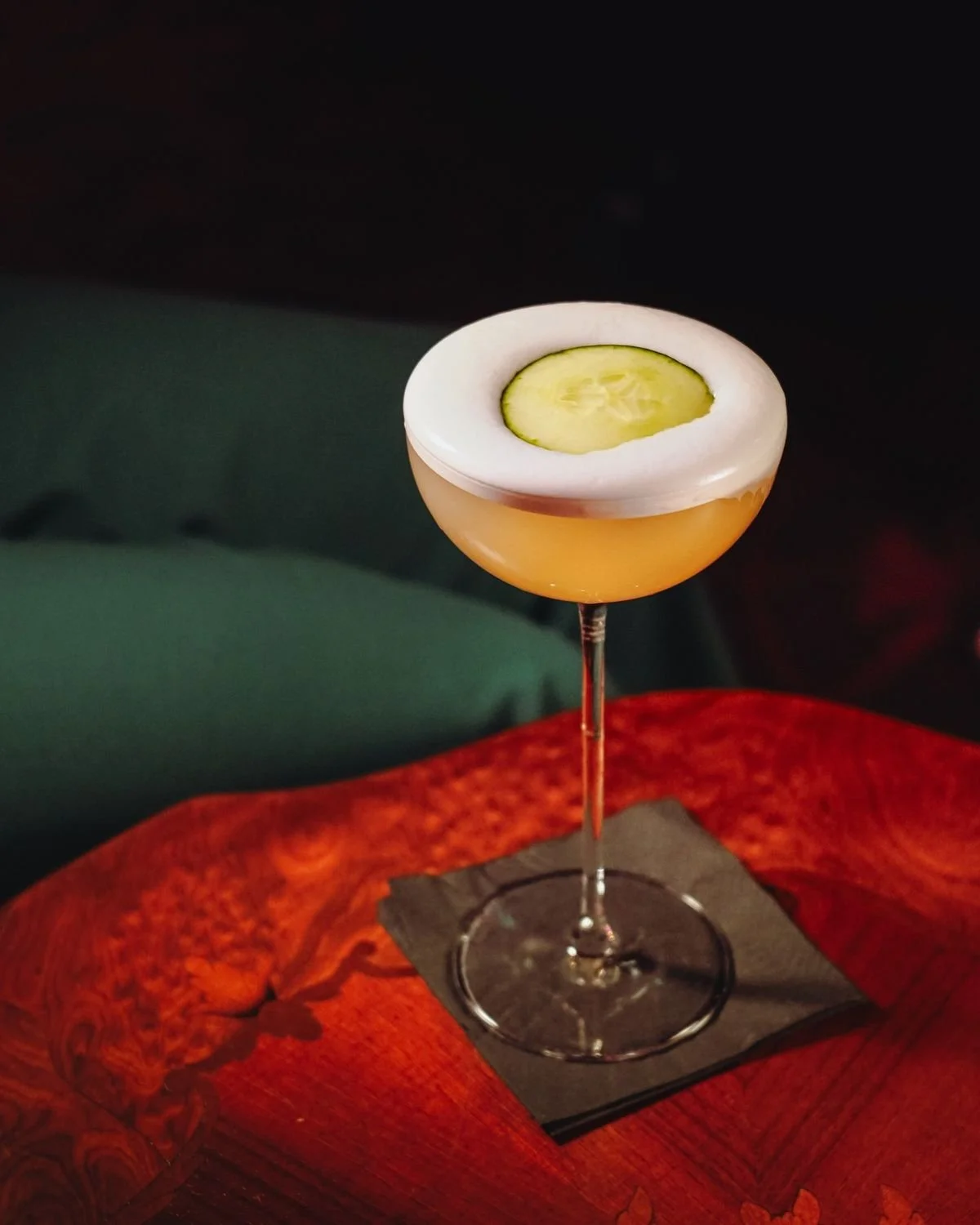 Cóctel en copa larga con rodaja de pepino y espuma blanca en la parte superior, sobre una superficie de madera