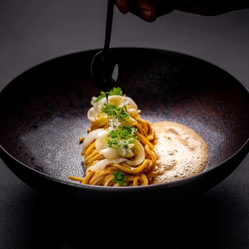 Persona rociando salsa sobre un plato de fideos con cebolla y cilantro en un cuenco negro.