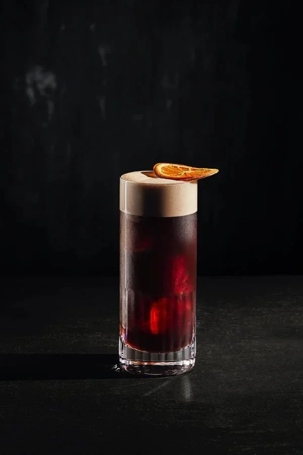 Copa de cóctel oscuro con espuma y una rodaja de naranja en la parte superior.