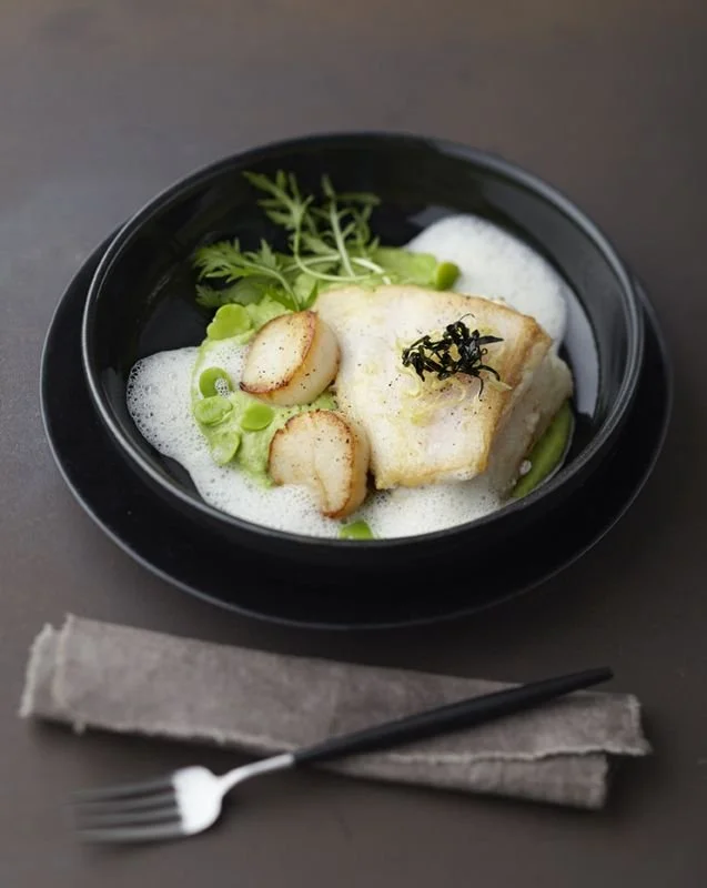 Plato de pescado con verduras y espuma, presentado en un bol negro con cubiertos y mantel de tela