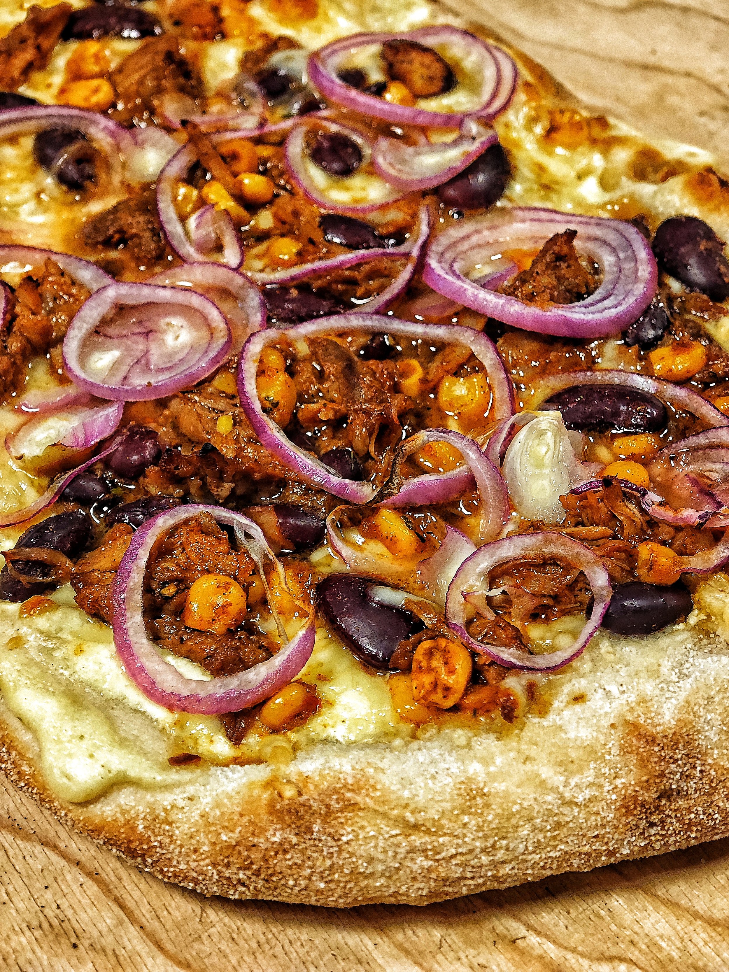 Pizza mit roten Zwiebelringen, schwarzen Bohnen, Mais und gewürztem Fleisch auf einem Holztisch.