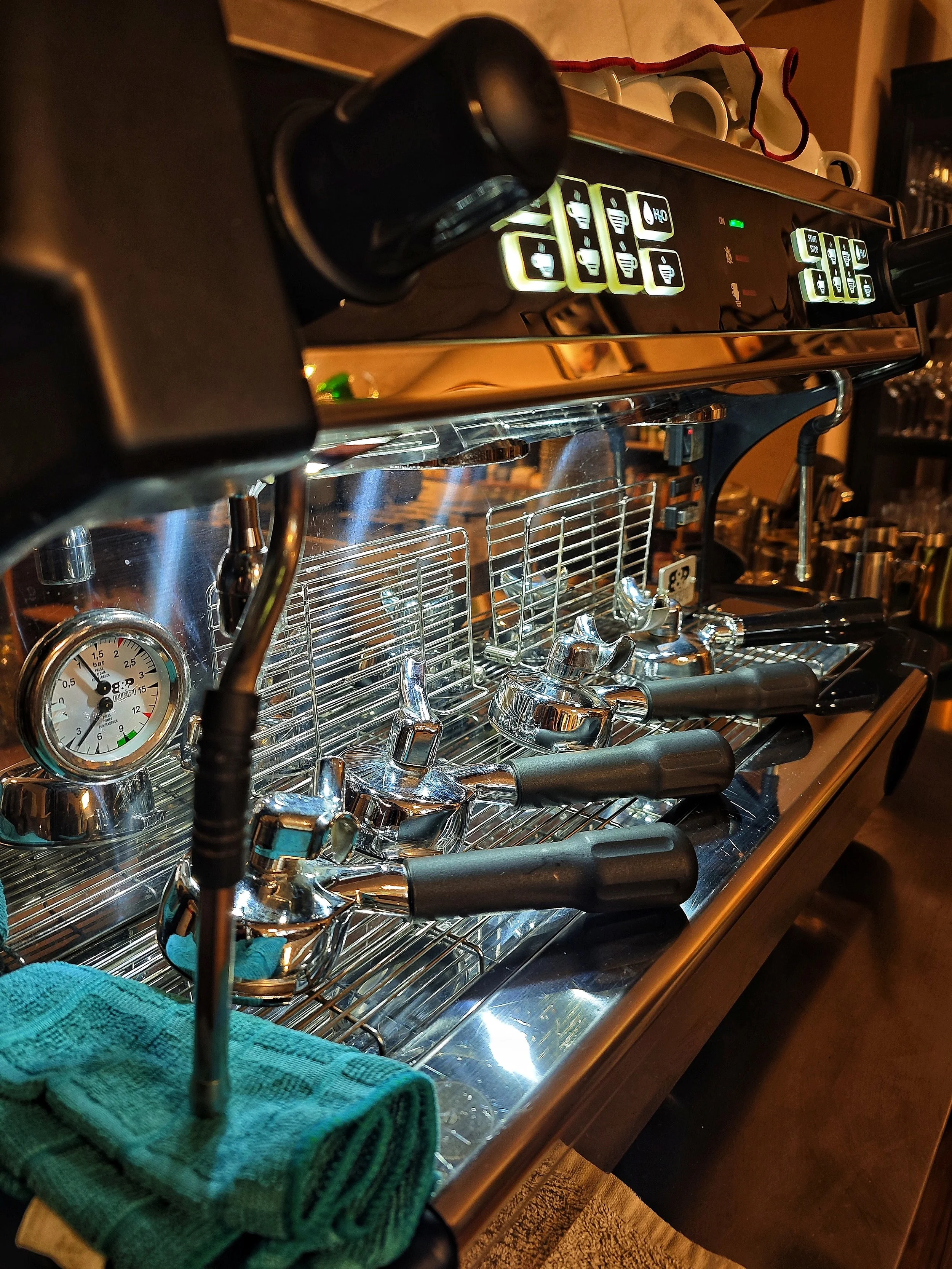 Eine Espressomaschine mit mehreren Espressoschläuchen und einem Druckmesser, dazu ein grünes Handtuch auf der Maschine.