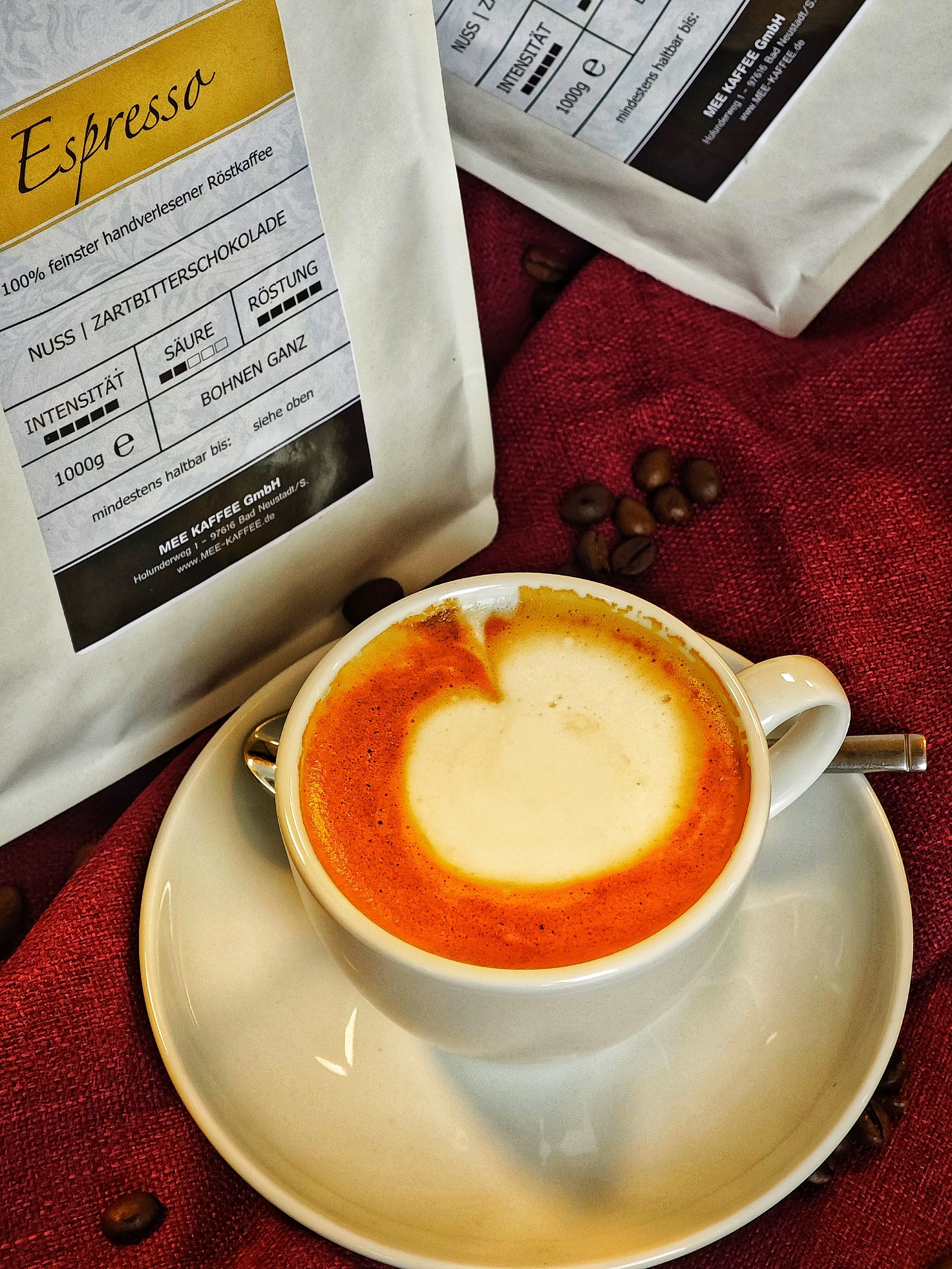 Cappuccino in einer weißen Tasse auf Untertasse, auf rotem Textilstoff mit Kaffeebohnen, Packung Kaffeebohnen und Kaffeeetikett im Hintergrund.