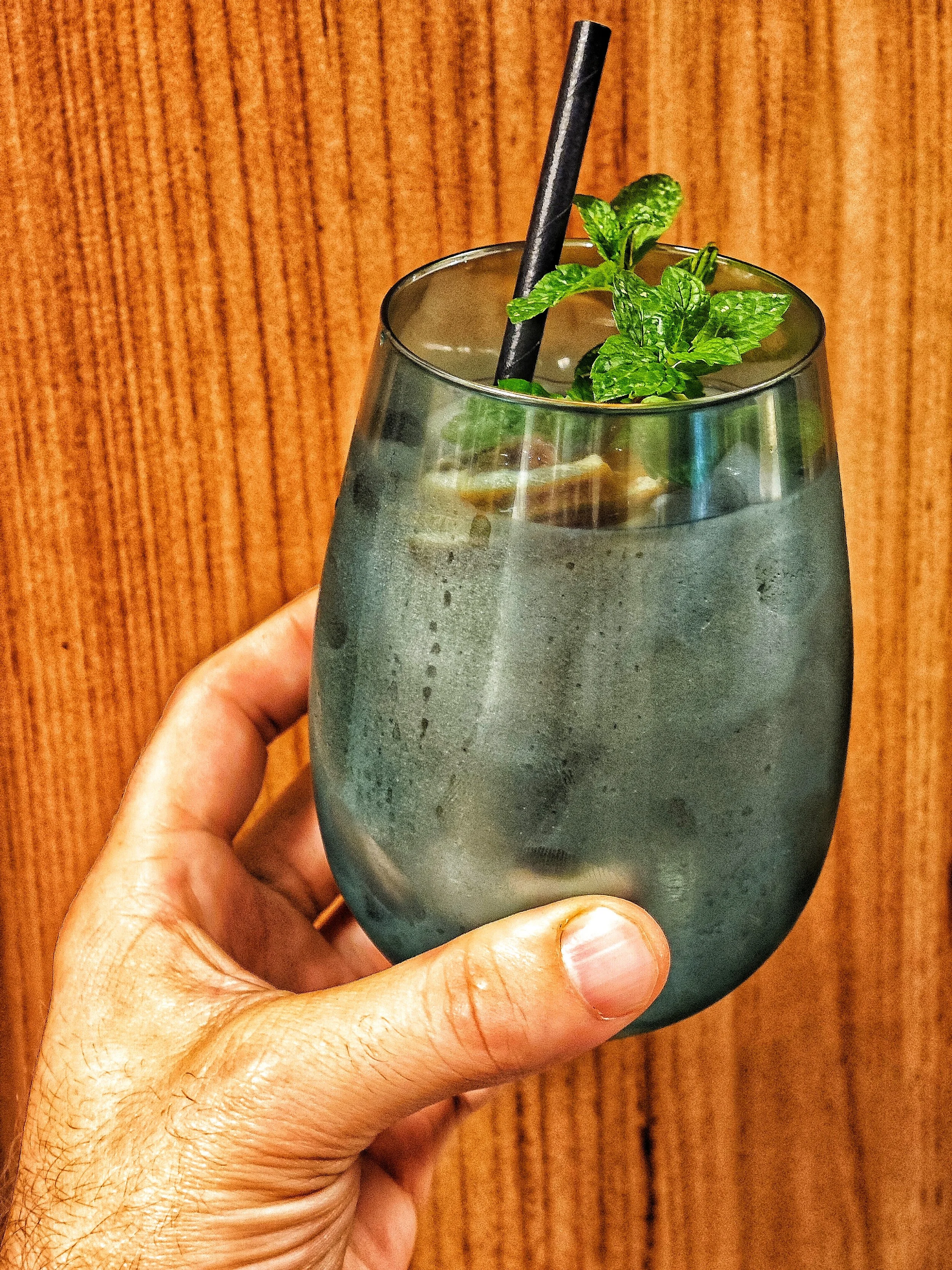 Hand hält ein Glas mit Wasser, Minzblättern, Zitrone und einem schwarzen Trinkhalm vor einem Holzhintergrund.