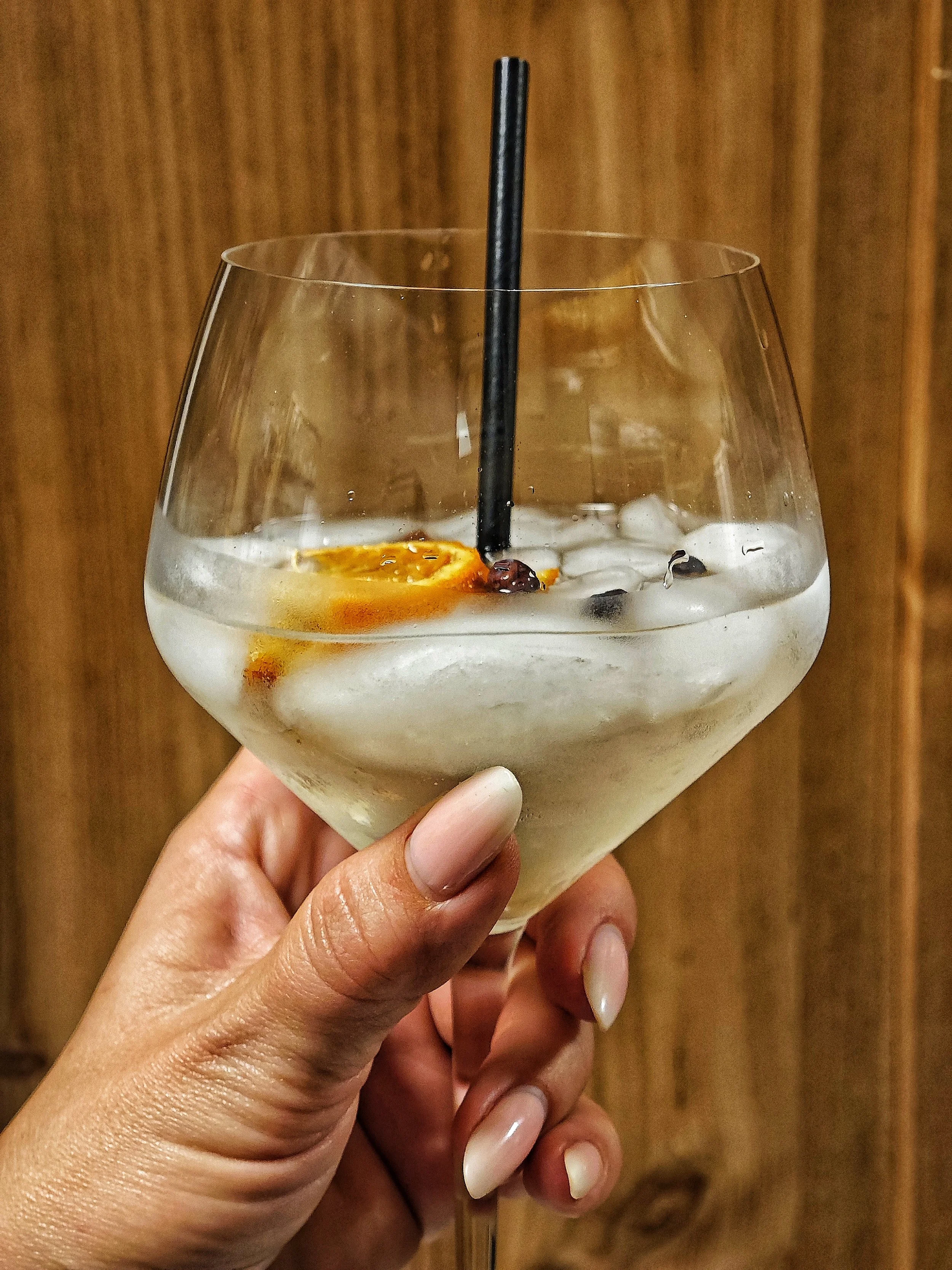 Ein Hand hält ein großes Cocktailglas mit Eiswürfeln, einem orangenen Fruchtstück und einem Strohhalm davor ein holzverkleideter Hintergrund.