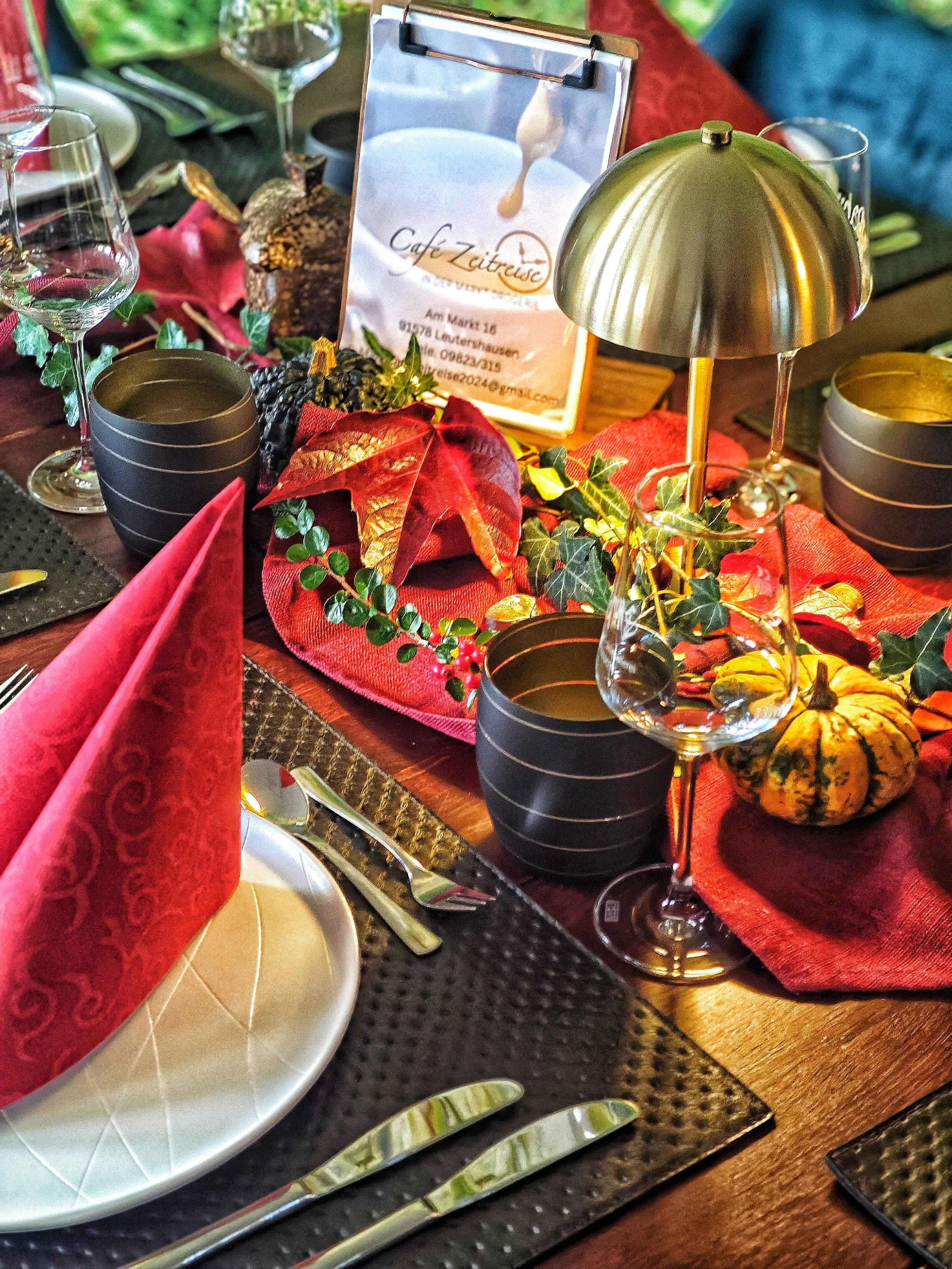 Ein festlich gedeckter Tisch mit roten Menagen, schwarz-golden Keramikbechern, Weingläsern, einem Menükarte und einer Herbstdekoration aus Blättern, Kastanien und Kürbissen, mit einer roten Tischdecke und dunklen Platzsets.