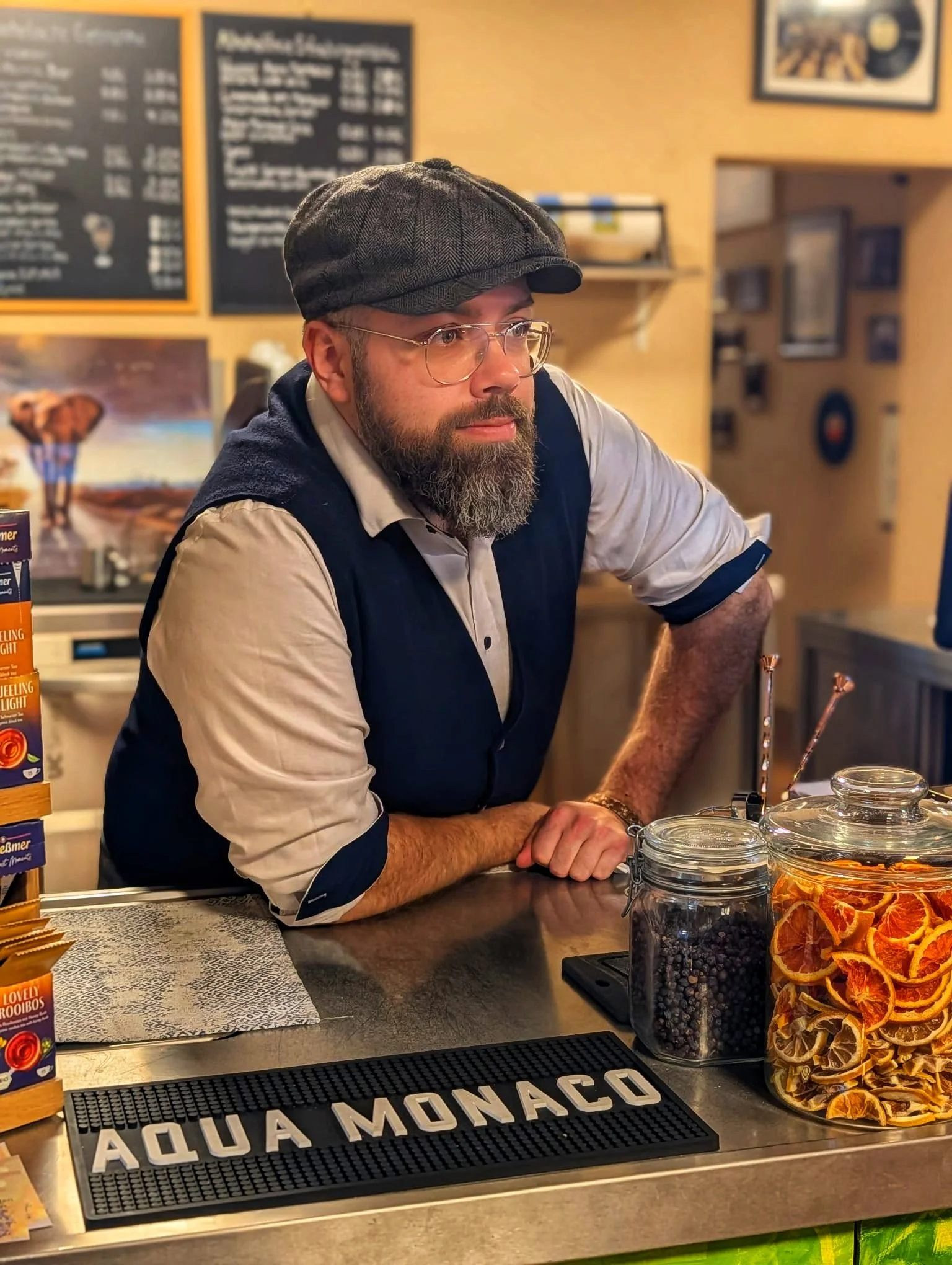 Ein Mann mit Brille und gestreiftem Cap hinter der Bar im Café, im Vordergrund stehen Gläser mit Orangen- und Beeren-Dekorationen sowie ein Zebrastreifen mit der Aufschrift 'Aqua Monaco'.