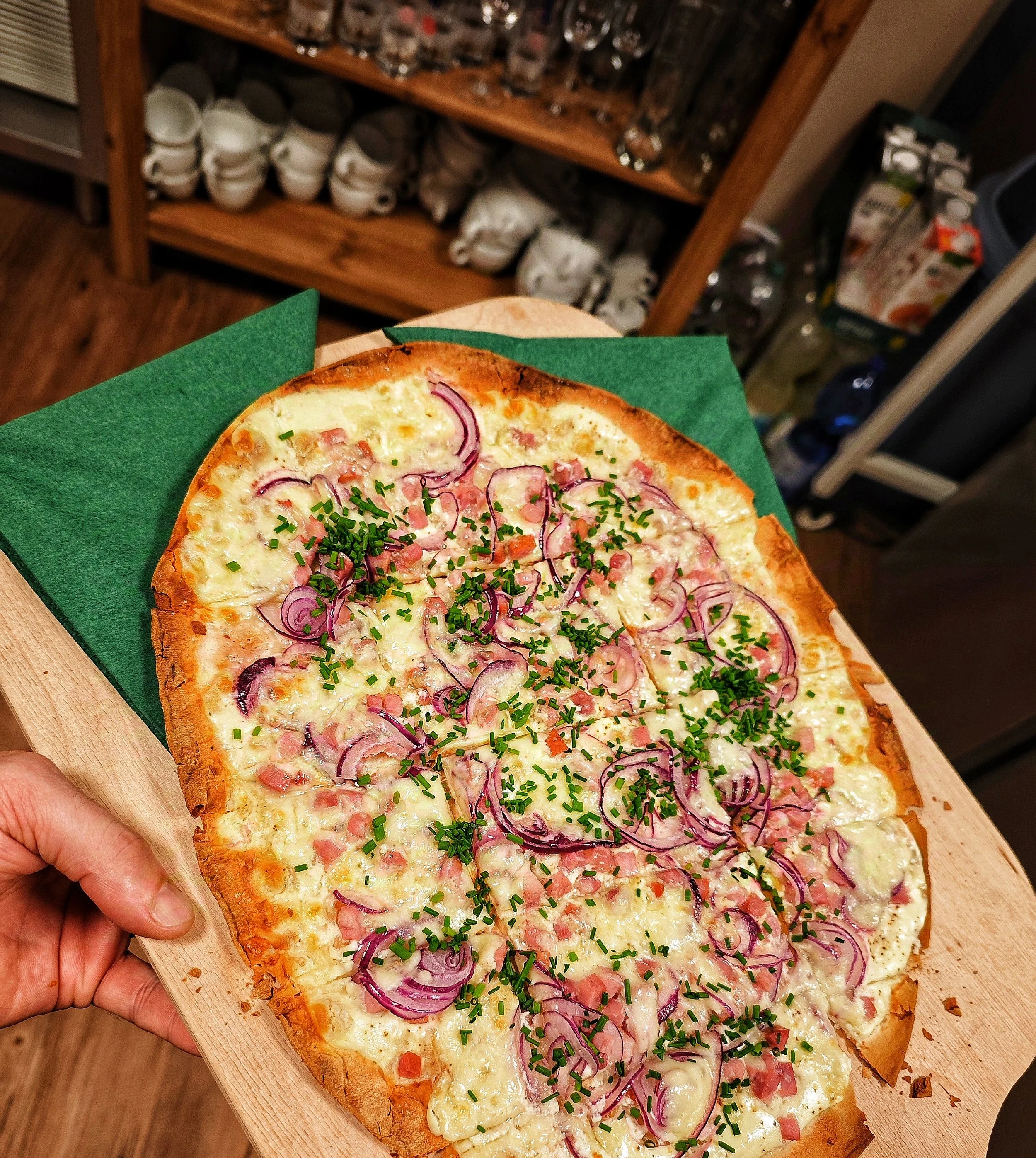Eine Flammkuchen mit Zwiebeln, Käse, und Kräutern auf einem Holzbrett, mit grüner Papierserviette darunter, im Hintergrund Regale mit Geschirr und Getränken.