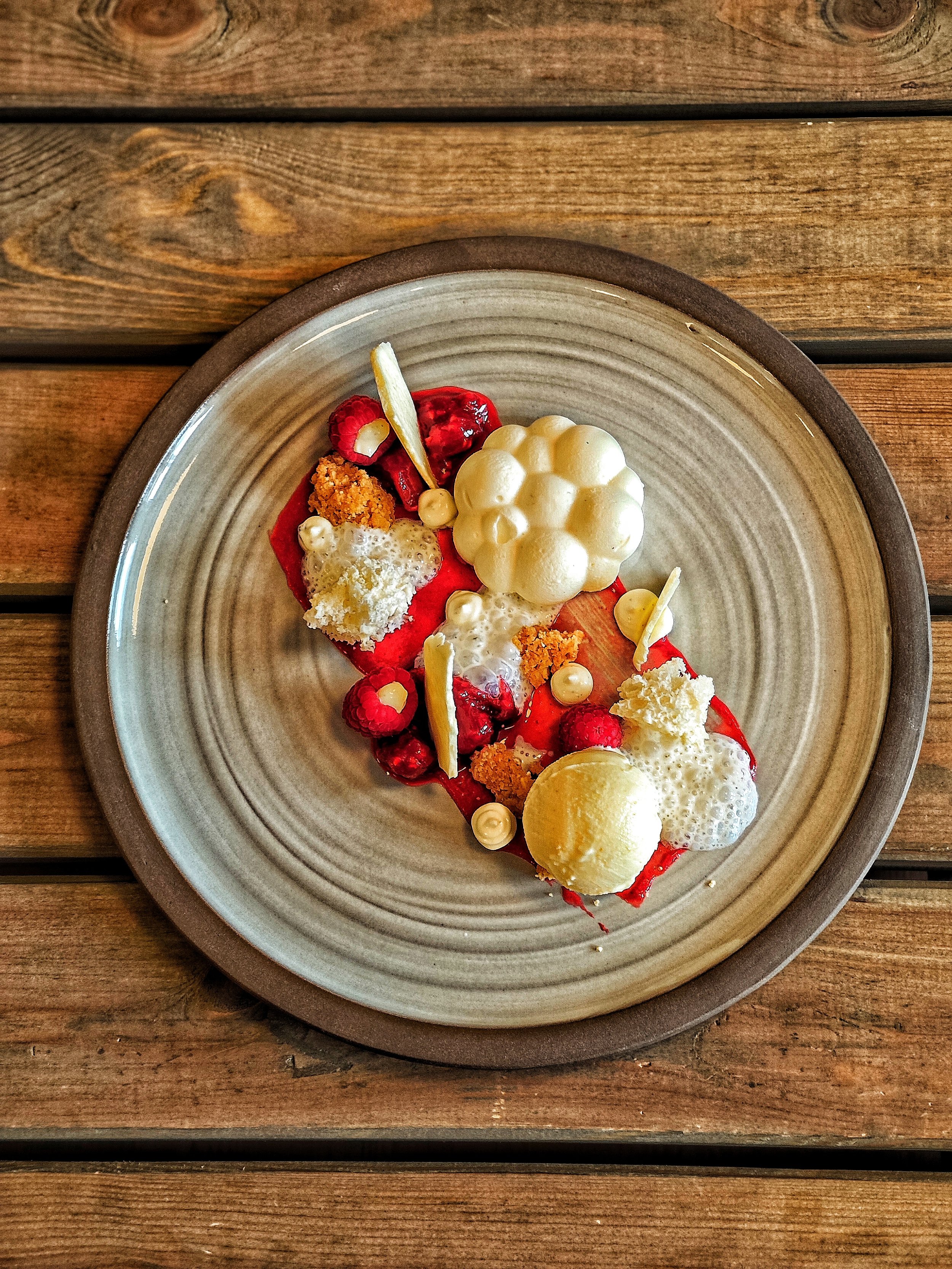 Ein Teller mit kreativer Dessertzubereitung, bestehend aus roten Früchten, weißen Schokoladenstücken, bunten Creme- oder Sandkapseln, weißen Bubbles, Gelée und verschiedenen Dekorationen auf einem Holztablett.