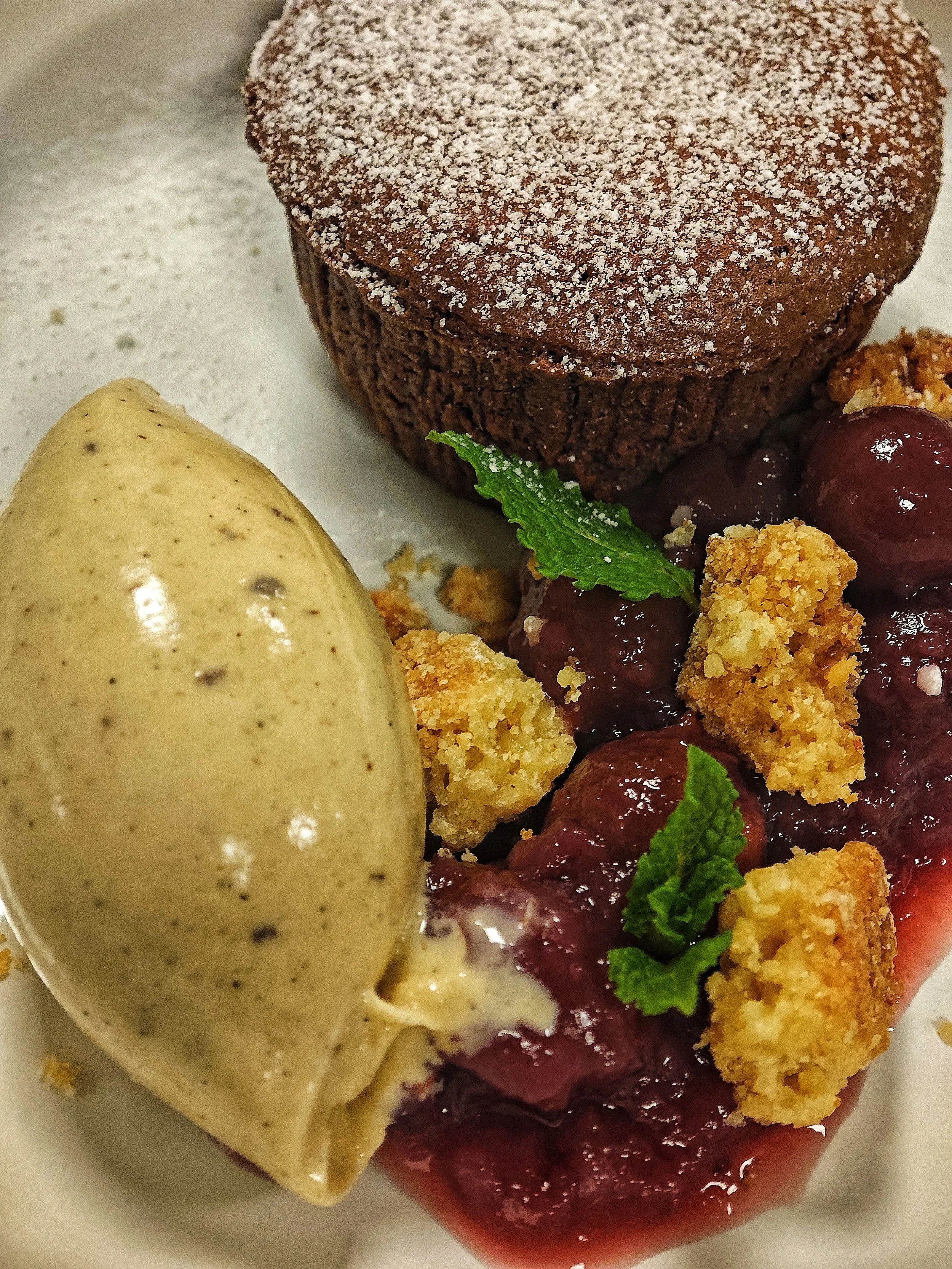 Auf einem Teller sind ein Schokoladenkuchen, eine Kugel Vanilleeis, Preiselbeerkompott und Streusel mit Minzblättern. Als Dekoration ist Staubzucker auf dem Kuchen.