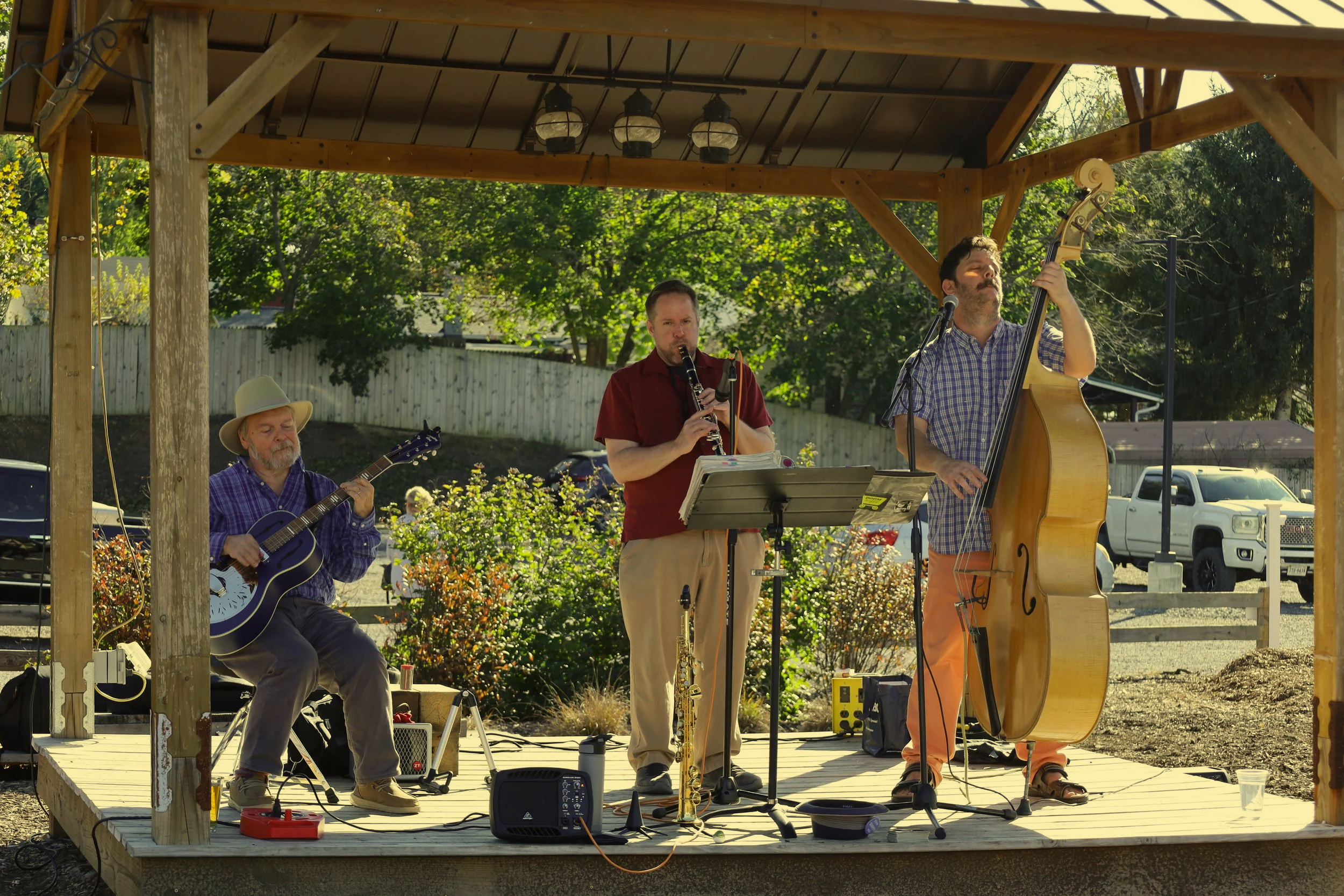 Three musicians performing on an outdoor stage, playing guitar, clarinet, and double bass, with a backyard and parked vehicles in the background.