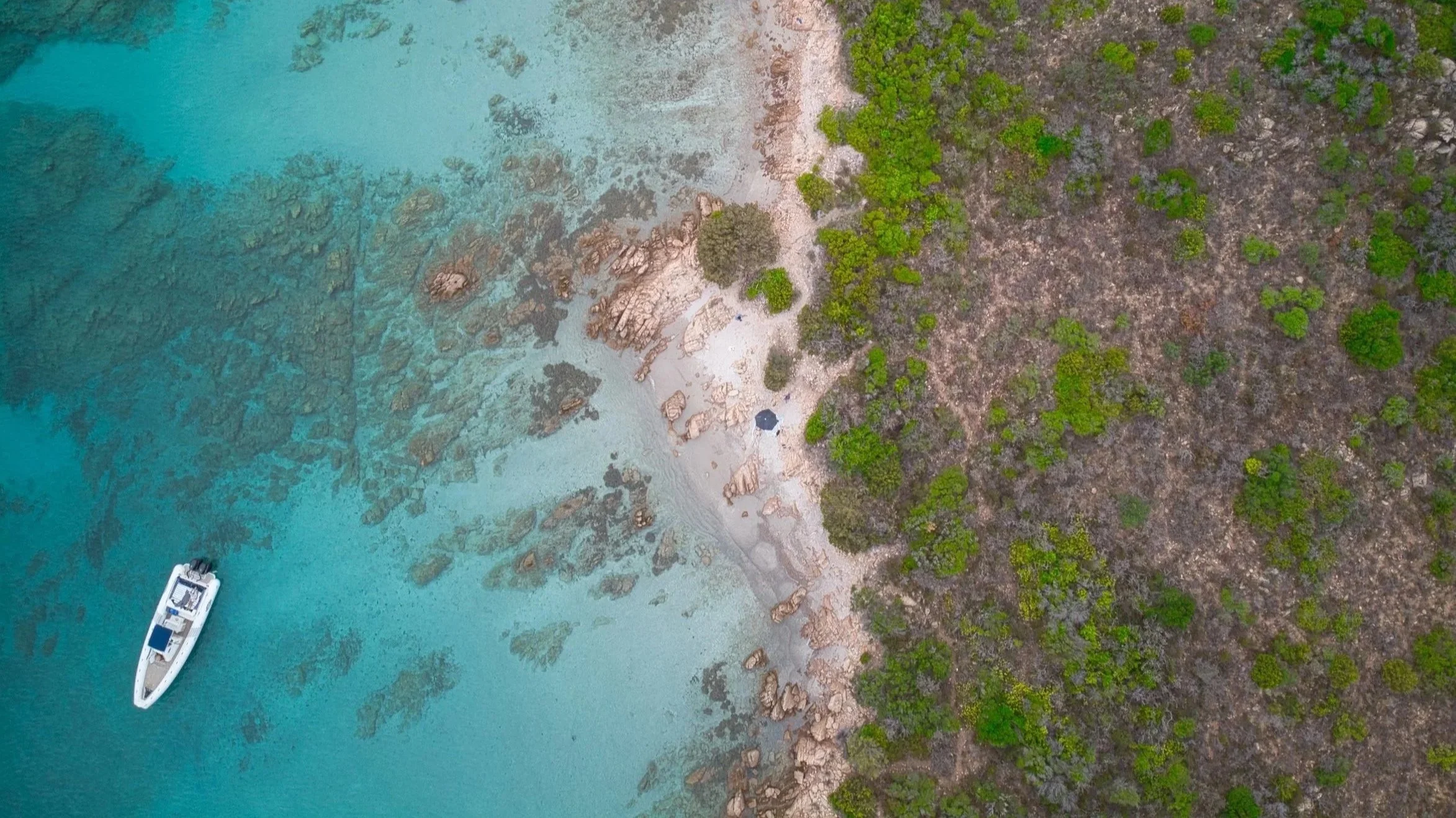 Vogelperspektive auf eine Bucht mit einem weißen Boot im klaren, blauen Wasser, umgeben von Felsen und einer Küste mit Wäldern.