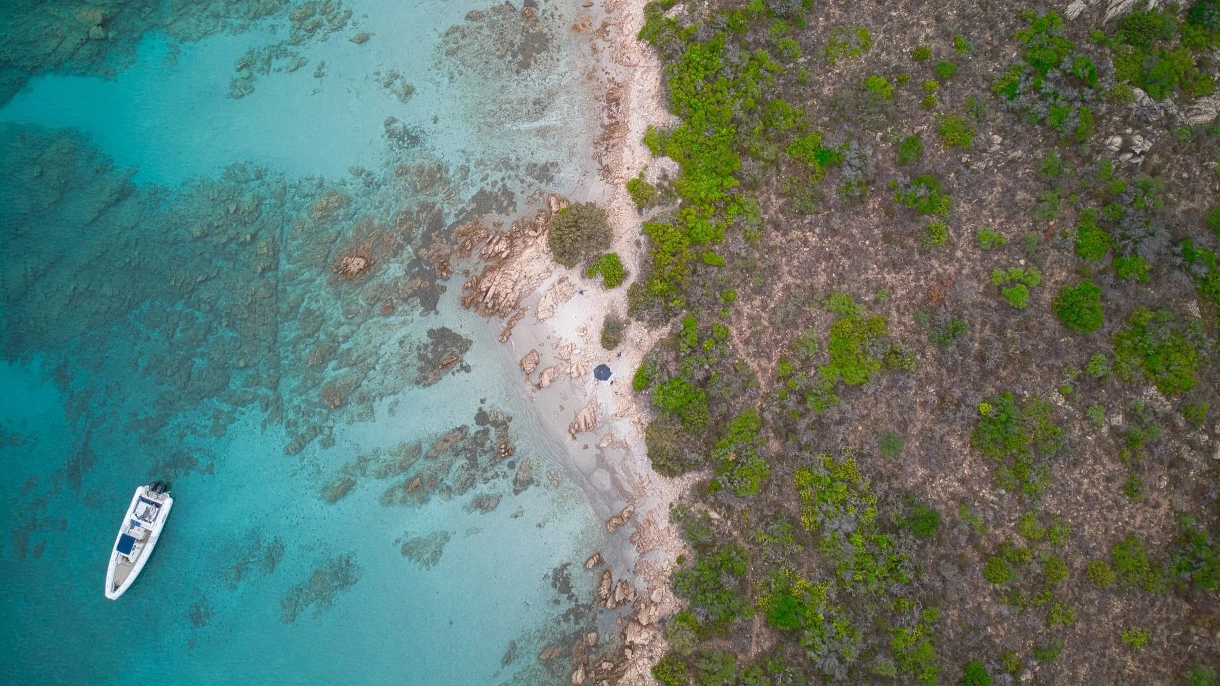 Vogelperspektive auf eine Küste mit Sandstrand, Felsen und grünem Bewuchs, sowie ein ins Wasser reichendes Segelboot